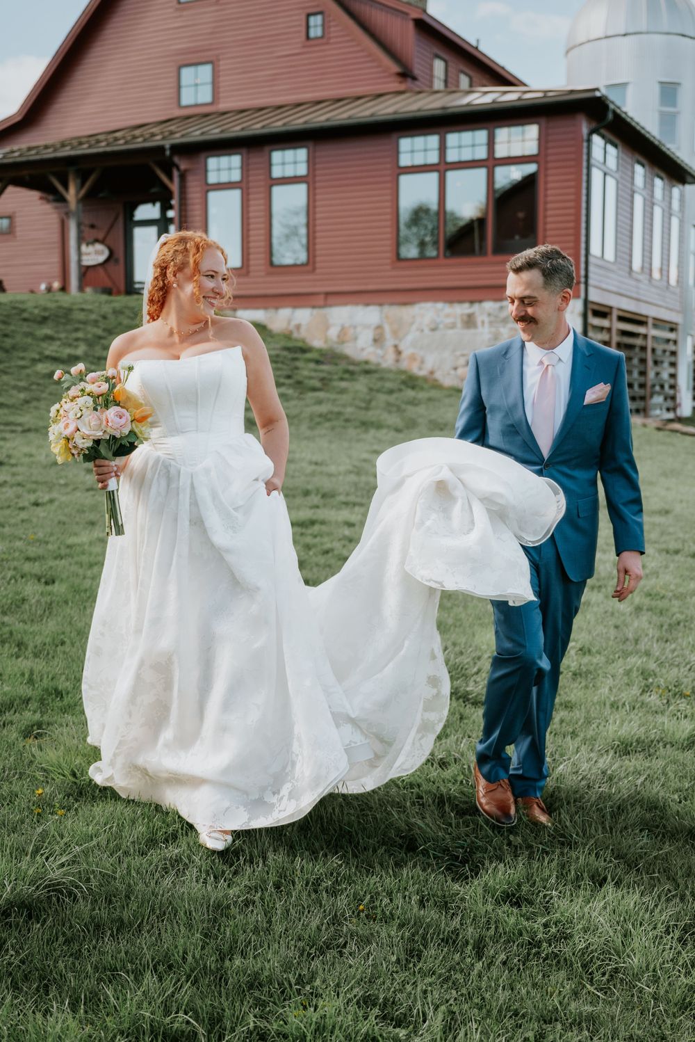 Bride and groom walking on lush green grass near a red barn venue, bride holding a colorful bouquet.
