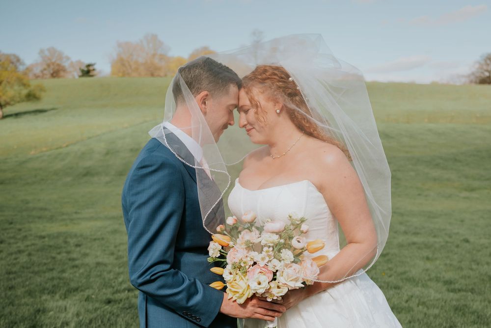 Bride and groom sharing an intimate moment under a flowing veil, holding a white and yellow bouquet.