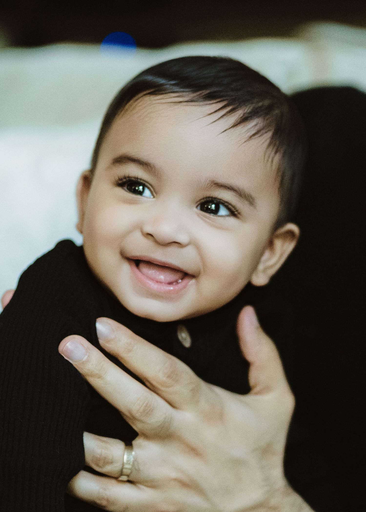 A smiling baby with dark hair and bright eyes being held by an adult wearing a silver ring.