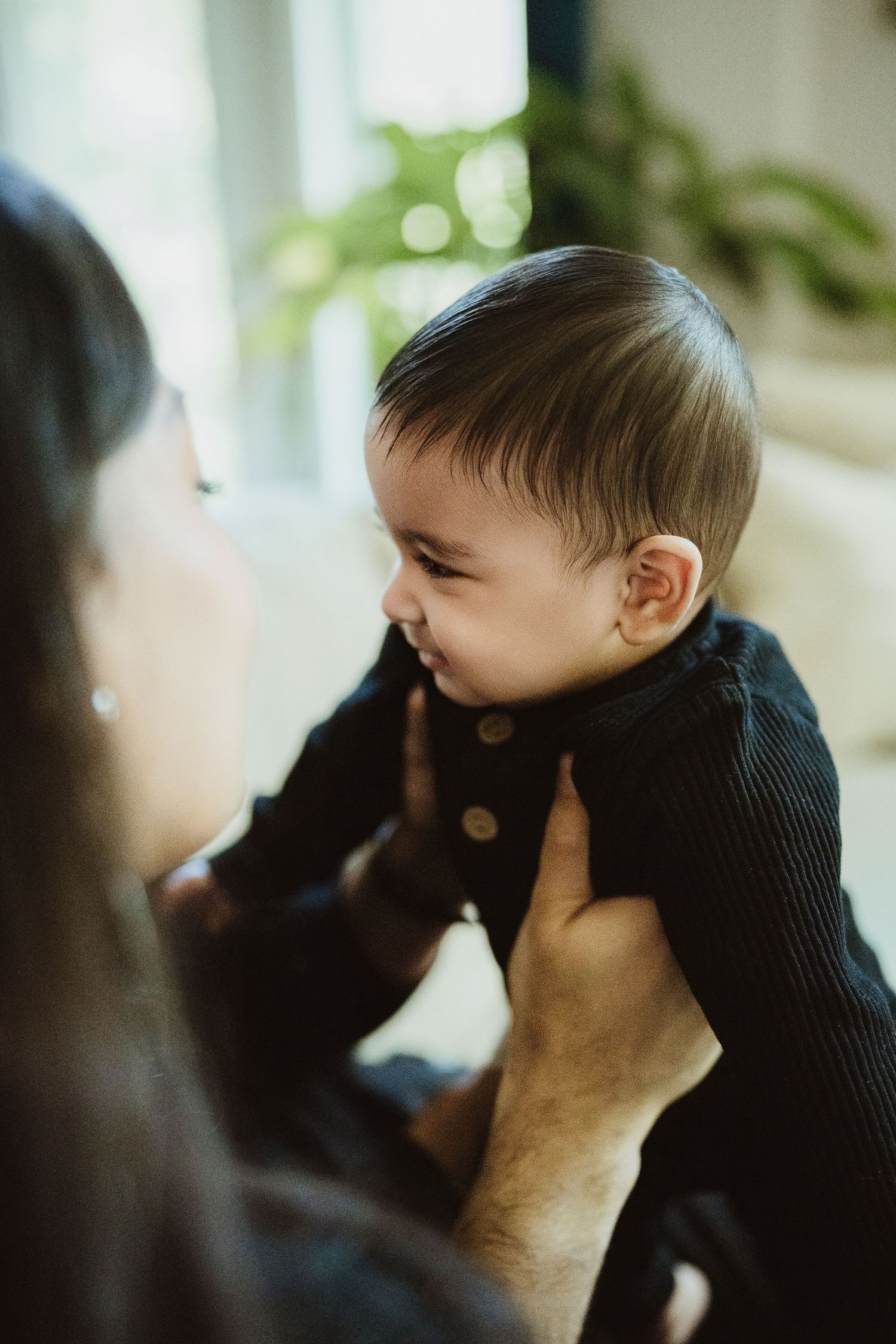 A smiling baby in a black knit outfit being held up by parents near a bright window.