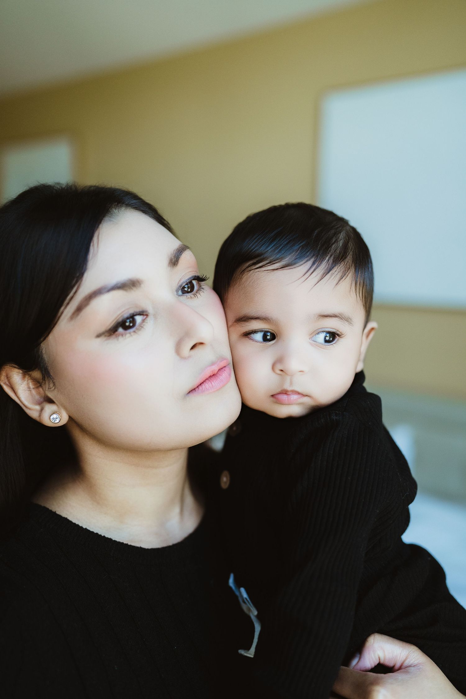 A young Asian mother in black holds her adorable dark-haired baby, both looking pensively at the camera indoors.