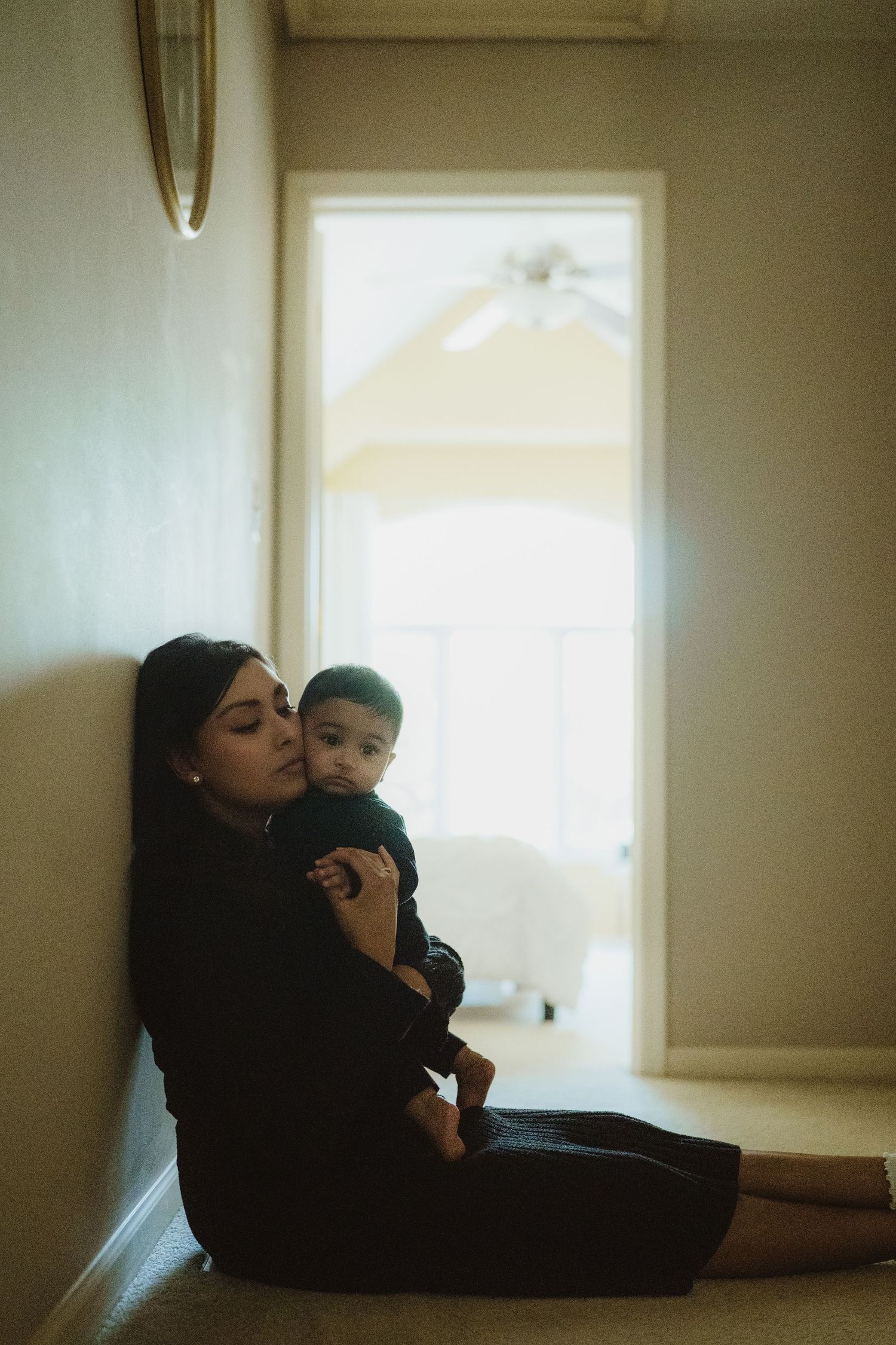 A young mother in black sits on the floor holding her baby, leaning against a wall in a softly lit hallway.