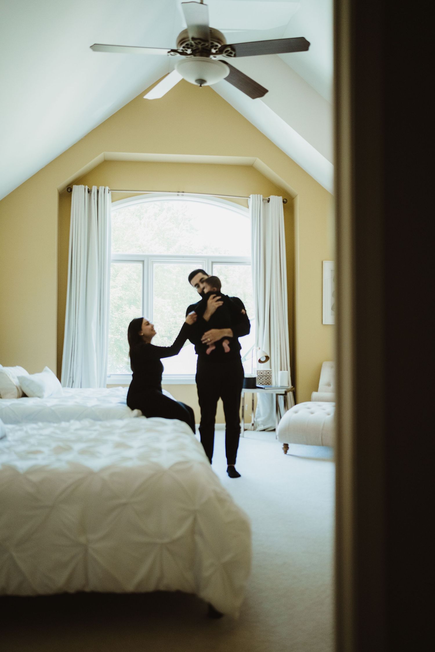 A family shares a joyful moment in a bright bedroom with vaulted ceilings, as a man holds a baby while a woman sits on the bed.
