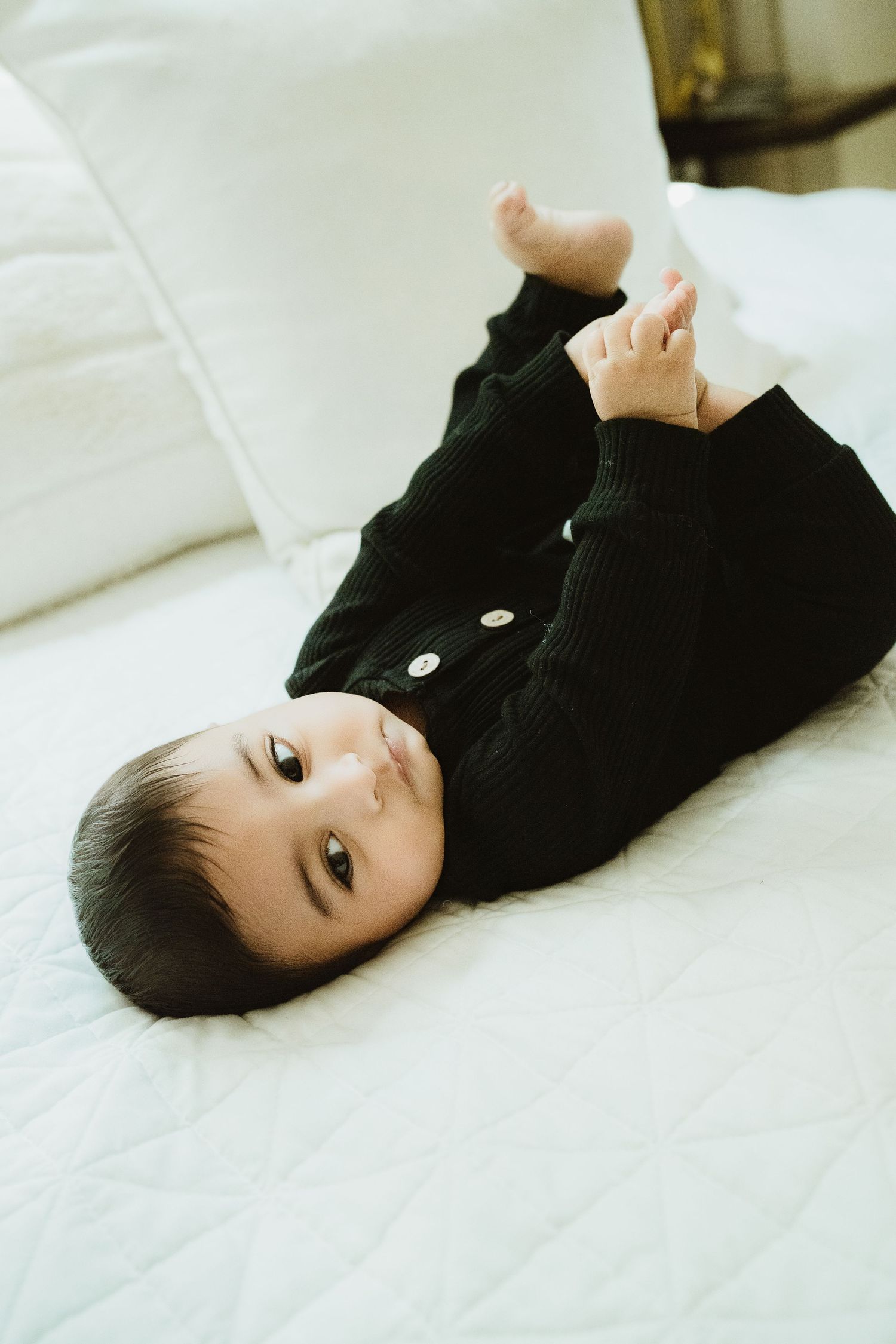 Baby in black knit outfit lying on white bed, looking at camera with bare feet raised.