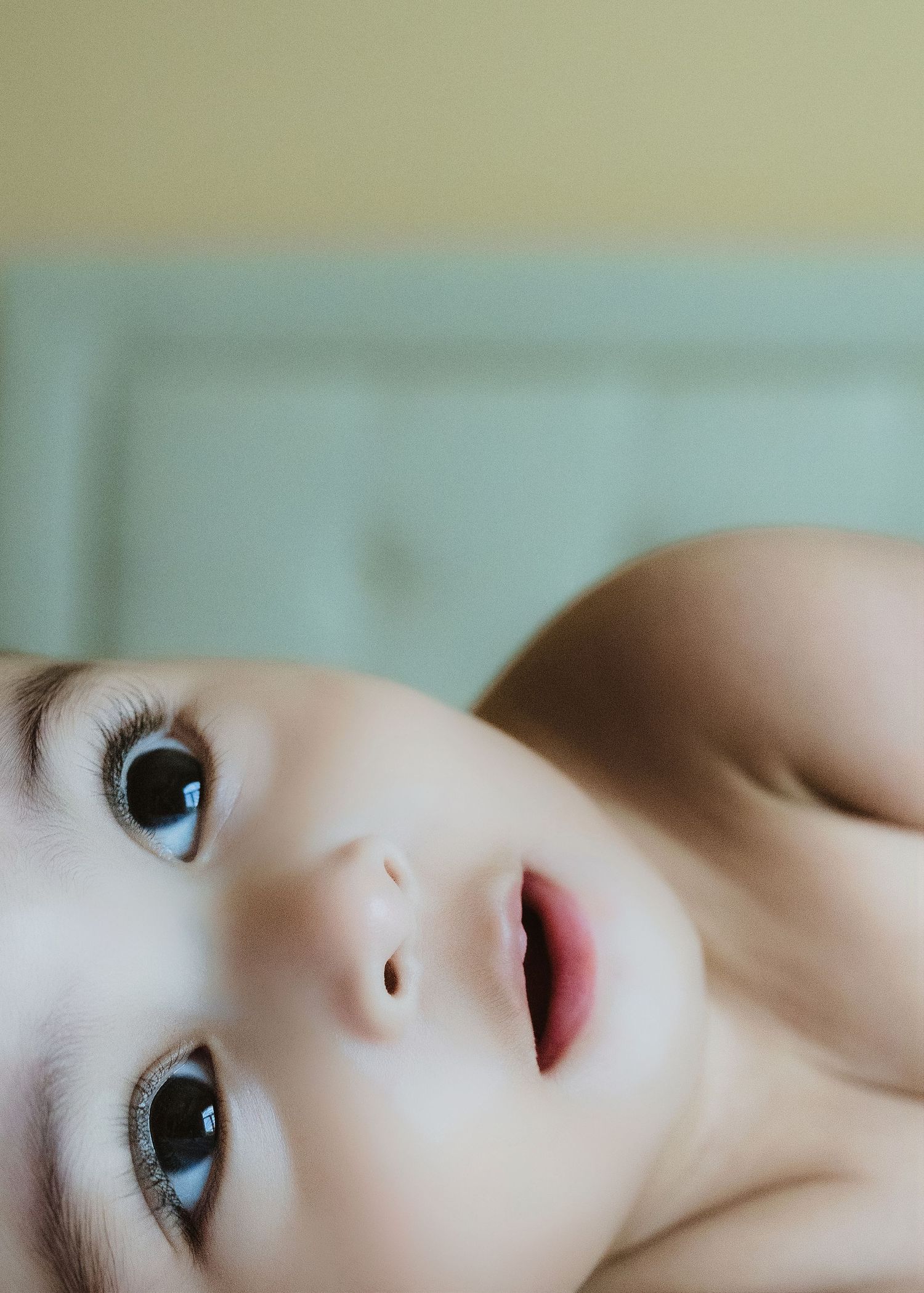 Close-up of a curious baby with big blue eyes lying on their back, looking upward with mouth slightly open.