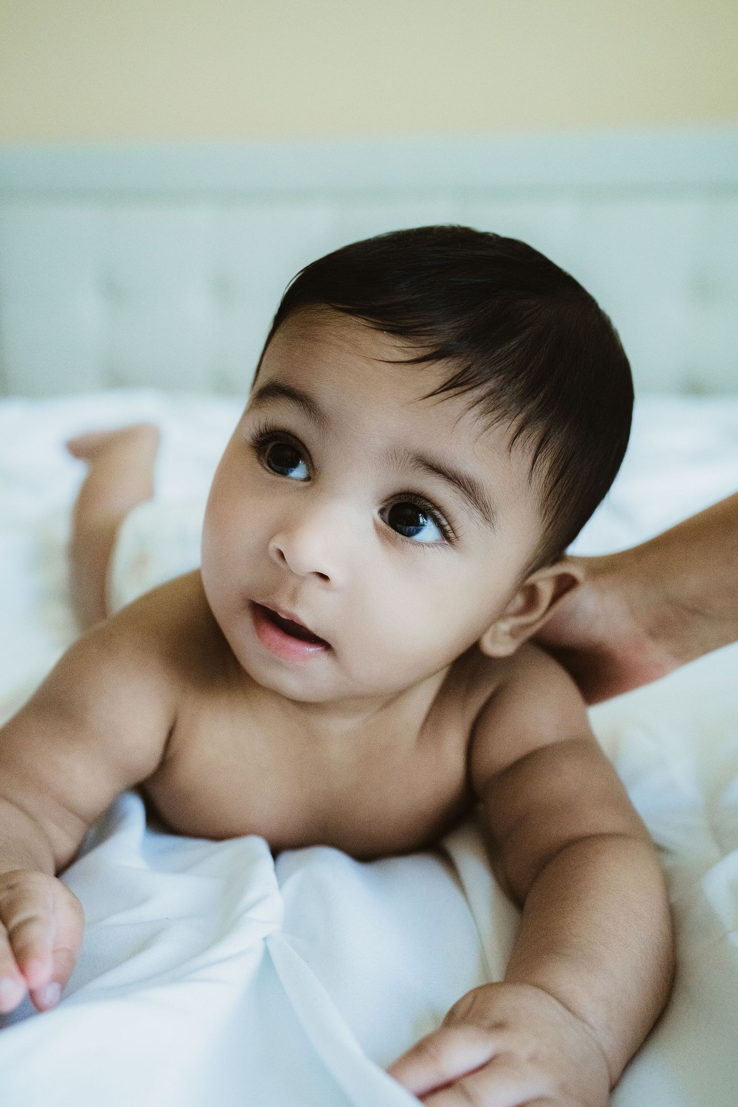 A cute baby with dark hair lying on a white bed, looking up with big brown eyes while an adult hand supports them.
