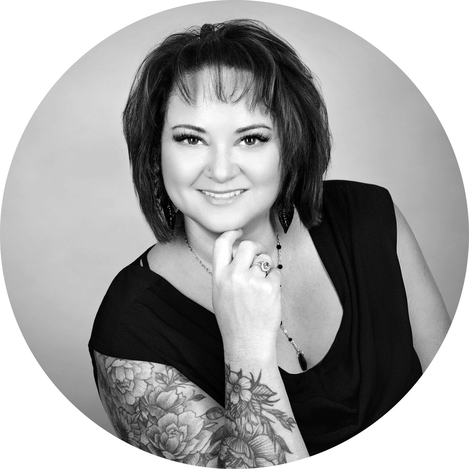 Black and white portrait of a smiling woman with tattoos on her arm, wearing a black top, posing with hand on chin.