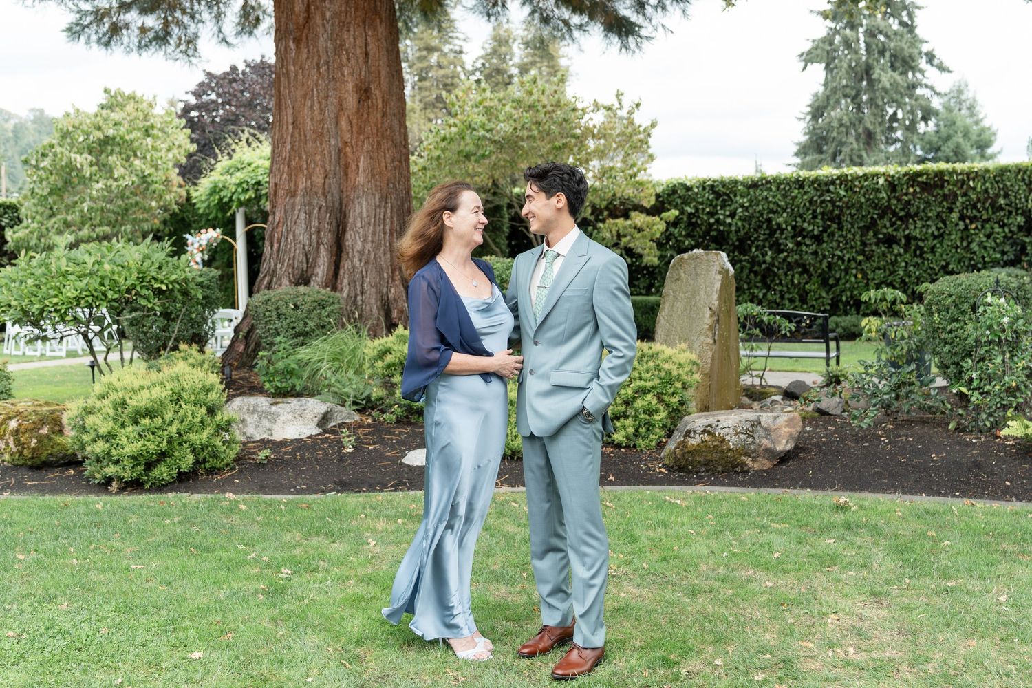 Couple in blue outfits holding hands and gazing at each other in a lush garden with tall trees.