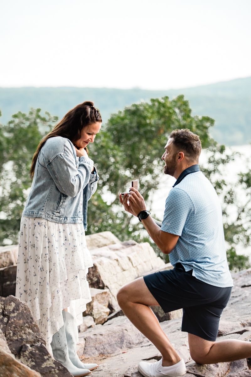 Man kneeling on rocky overlook proposing to emotional woman with ring box, scenic lake and trees in background.