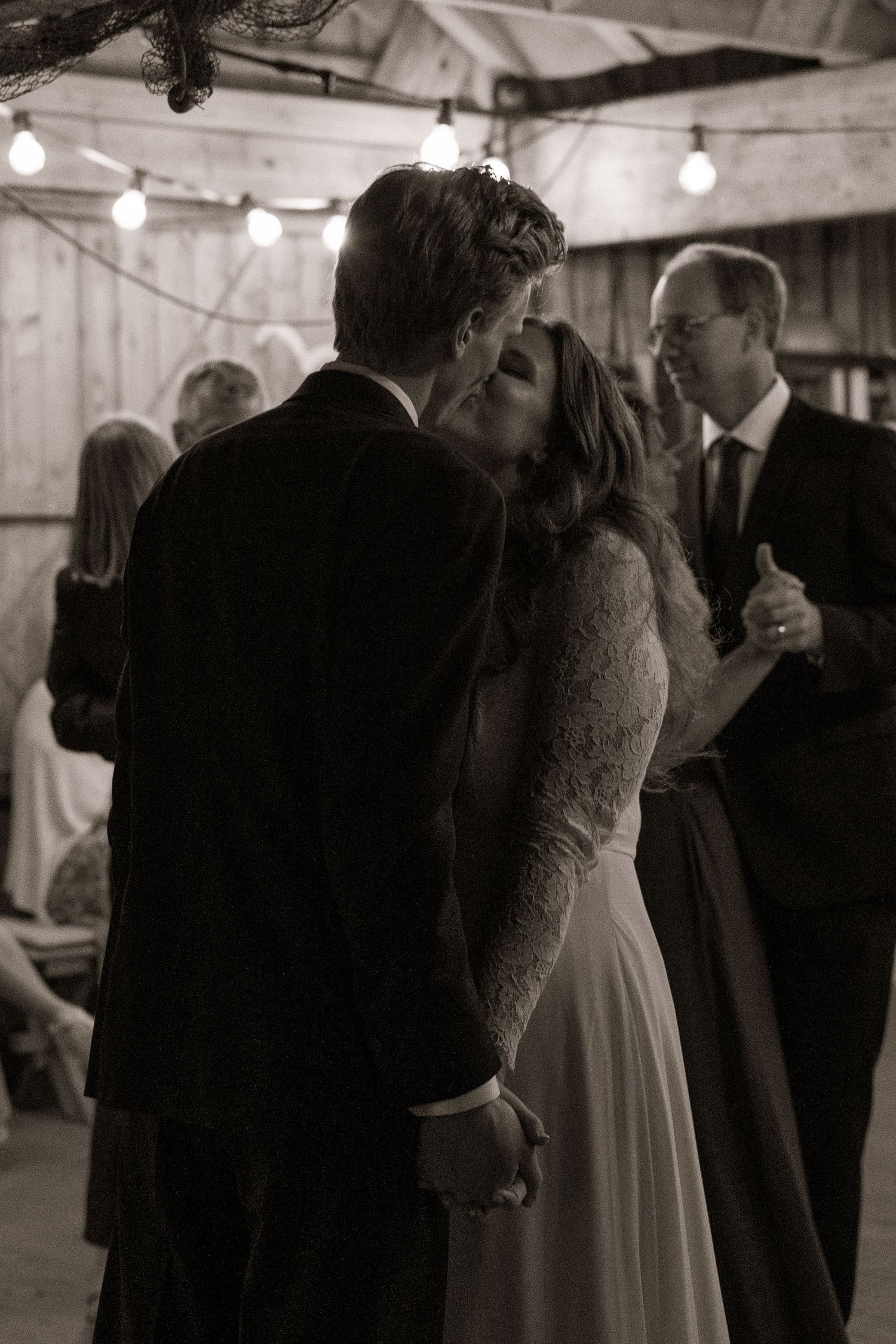 Black and white photo of a bride and groom sharing their first kiss at a rustic barn wedding reception.