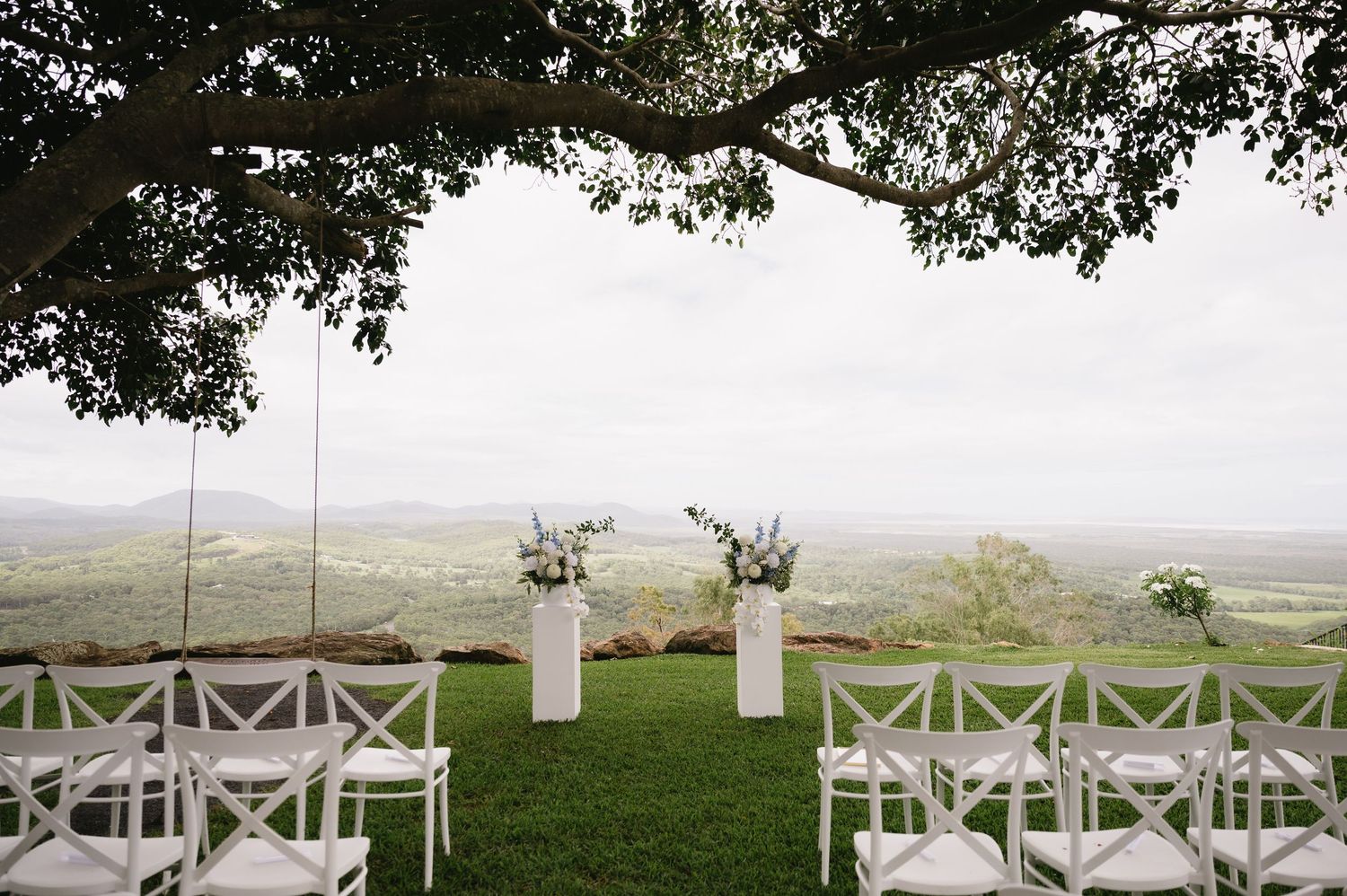 Elegant outdoor wedding ceremony setup with white chairs and floral arrangements overlooking a scenic valley landscape.