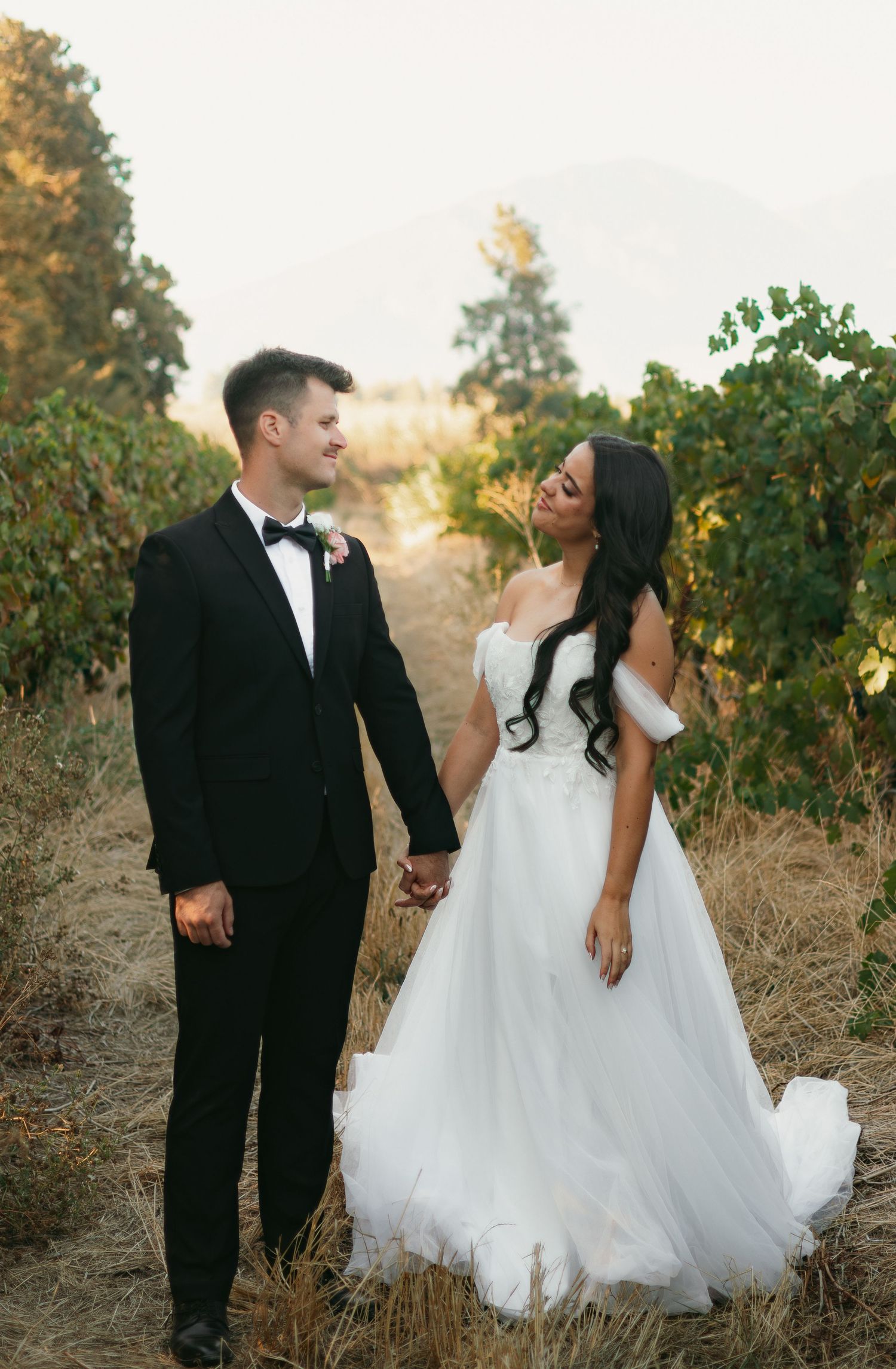 Bride in off-shoulder white gown and groom in black tuxedo holding hands in vineyard.