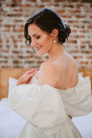 Bride adjusting her dress sleeve while standing near a brick wall.