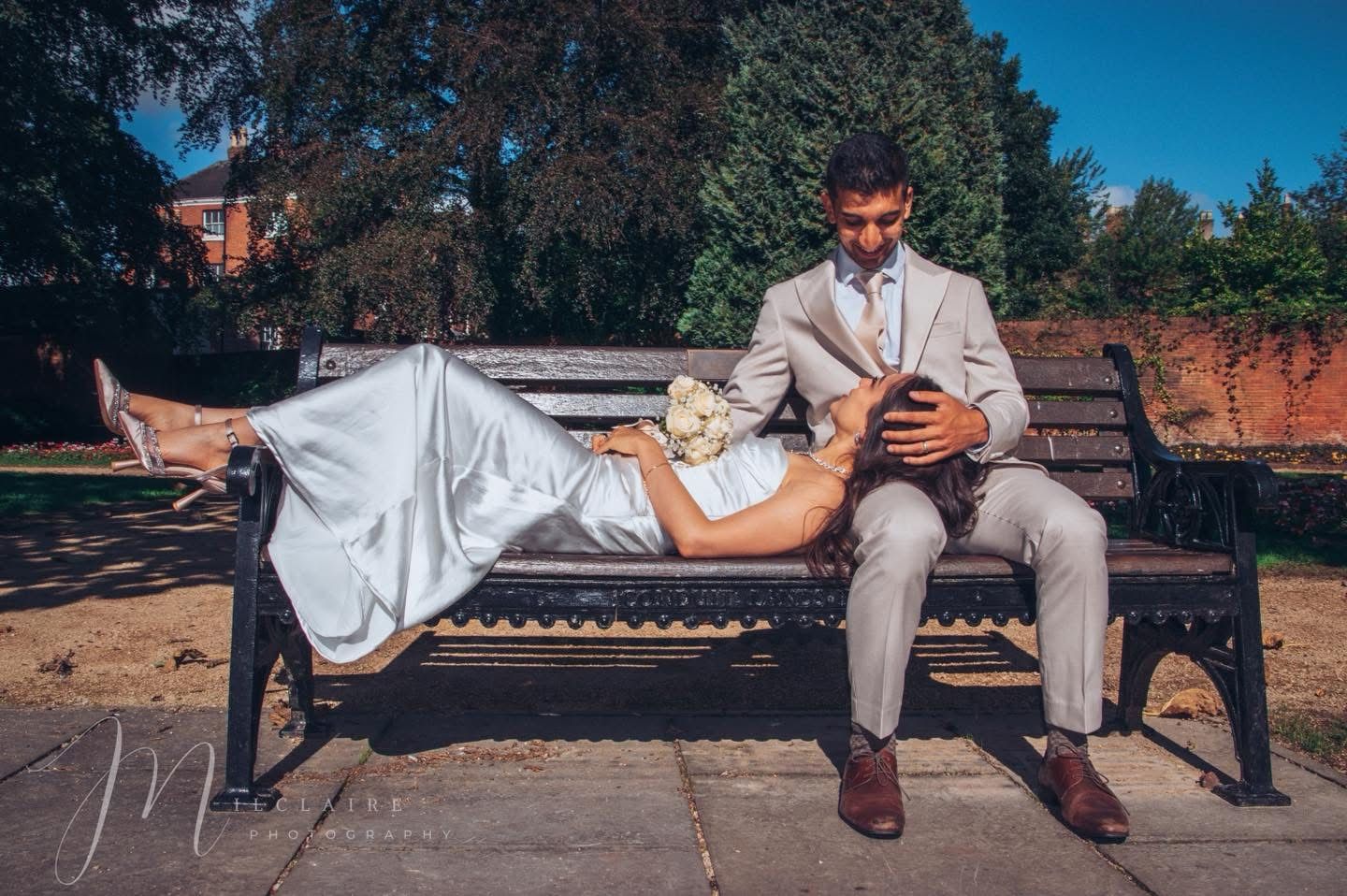 Bride in white gown lying on park bench while groom in gray suit looks at phone on sunny day.