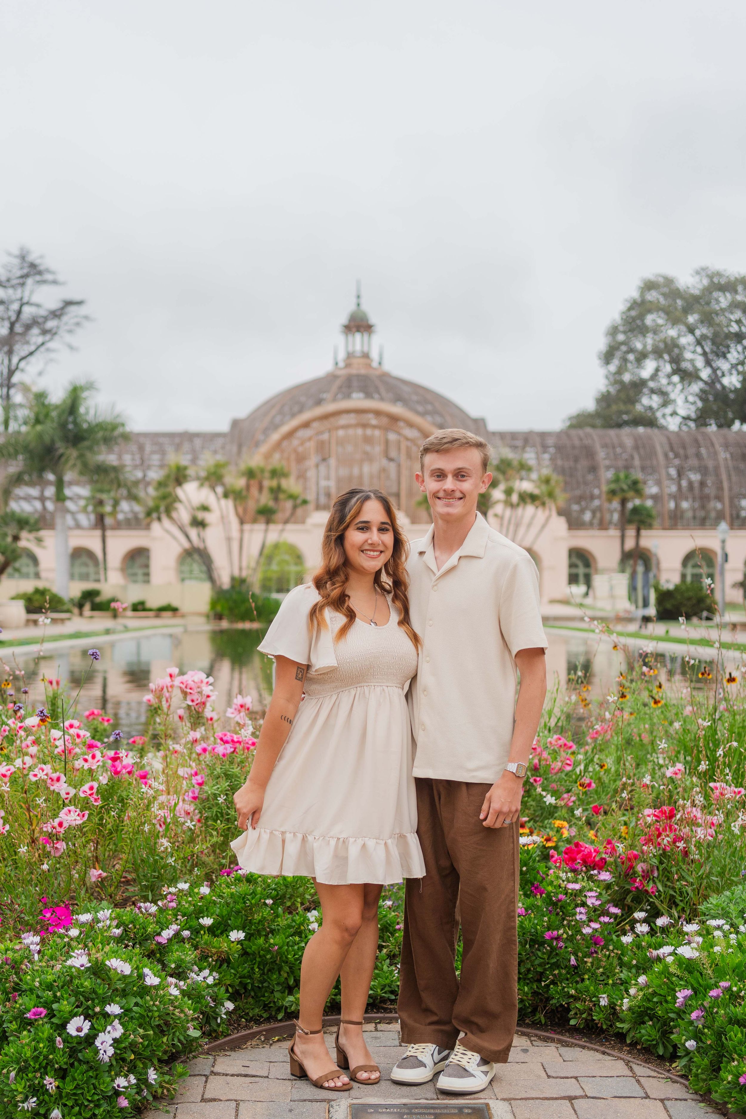 Couple poses together in front of blooming garden and historic building architecture.