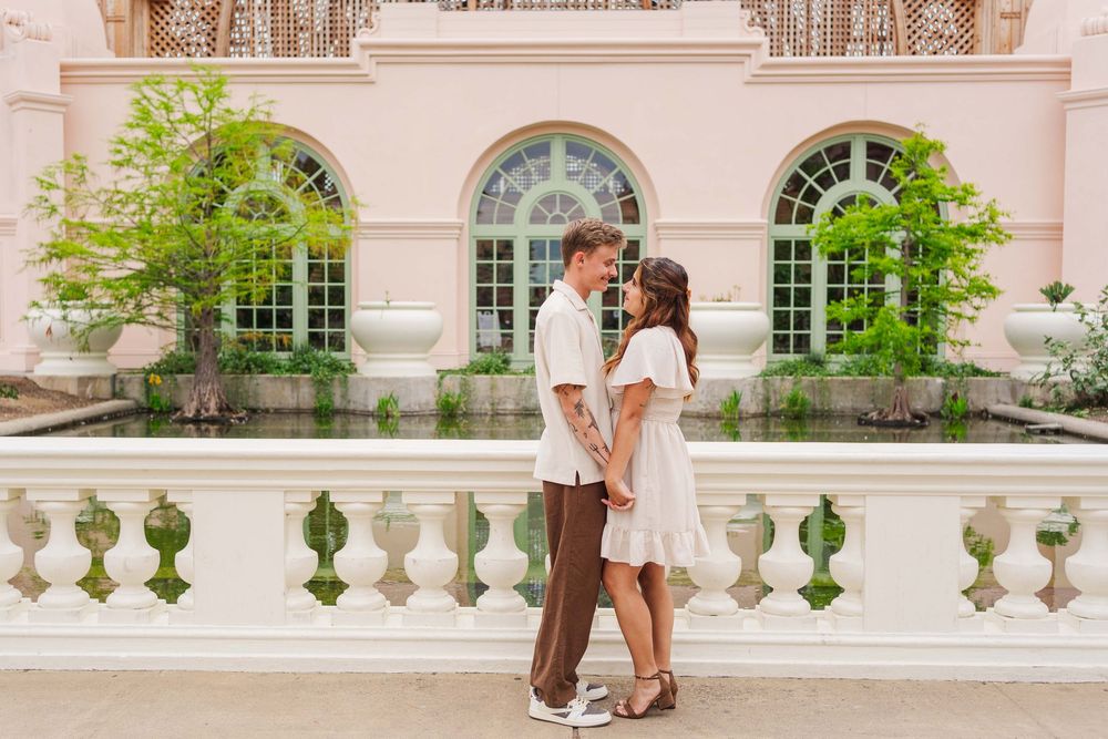 A couple stands close together on an elegant stone balcony with arched windows and potted plants in the background.
