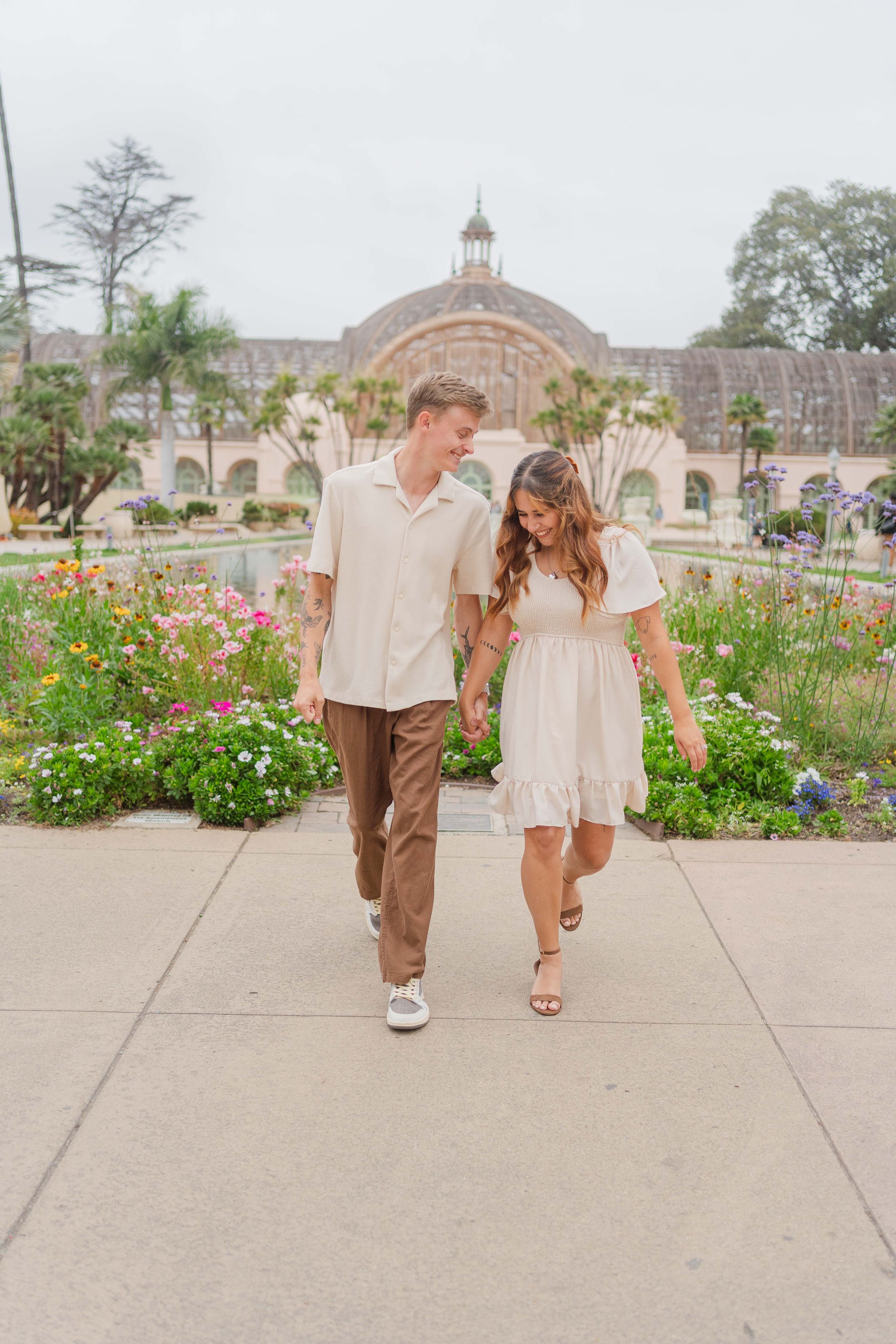 Two people walking hand in hand along a concrete path with colorful flowers blooming in the garden behind them.
