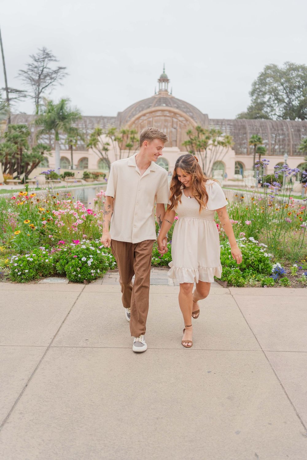 Two people walking hand in hand along a concrete path with colorful flowers blooming in the garden behind them.