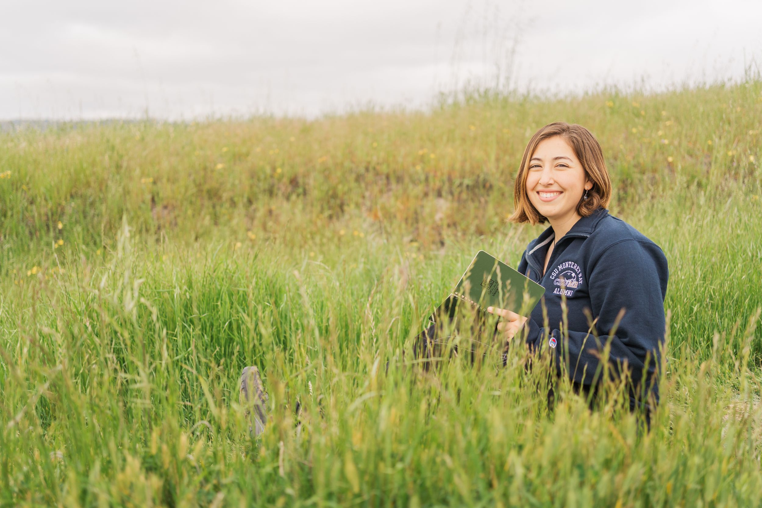 Someone wearing a dark blue sweater sits in a field of tall grass and wildflowers on a bright day.