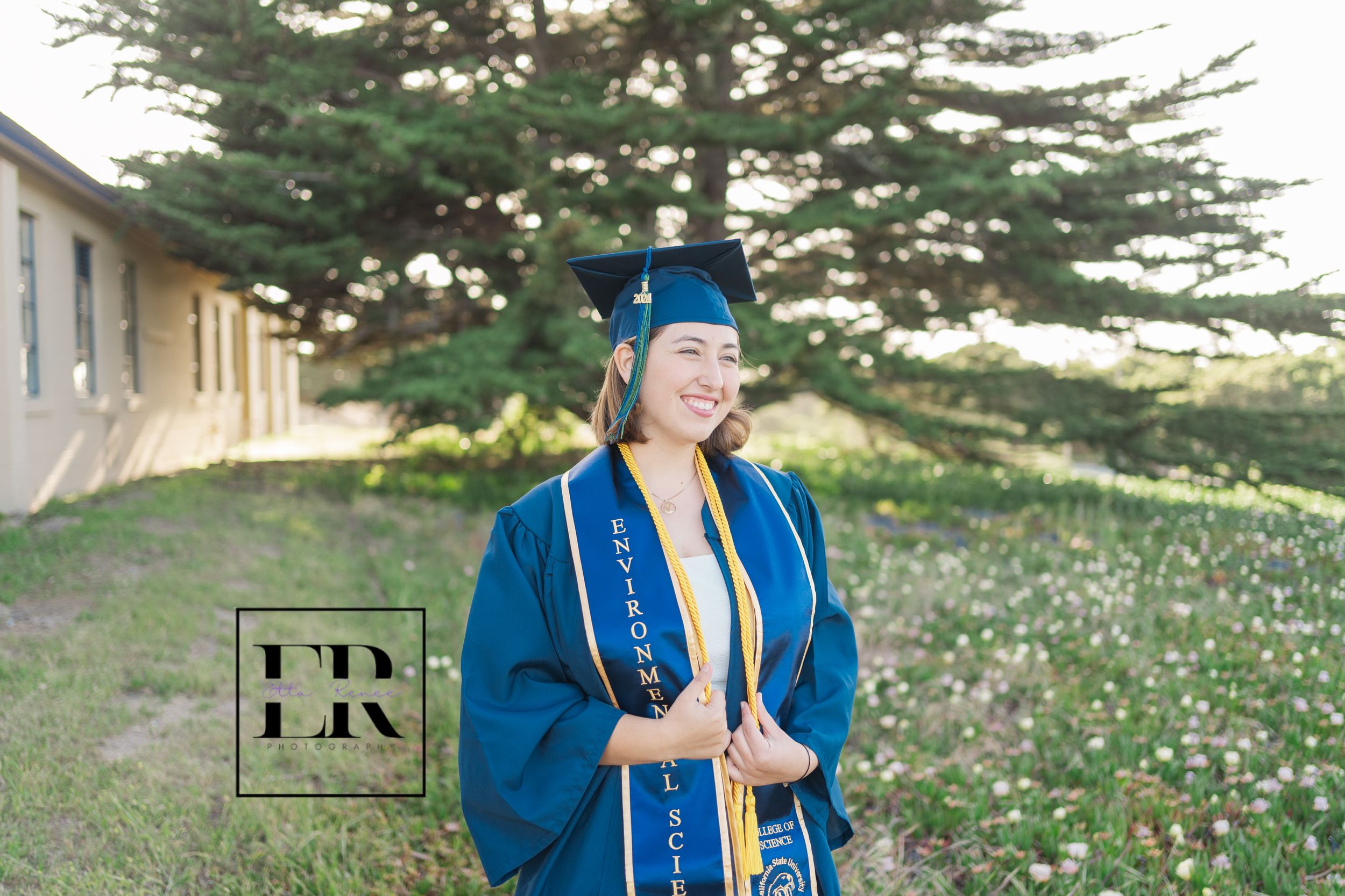 Graduate in blue cap and gown standing outdoors in front of evergreen tree and building on sunny day.