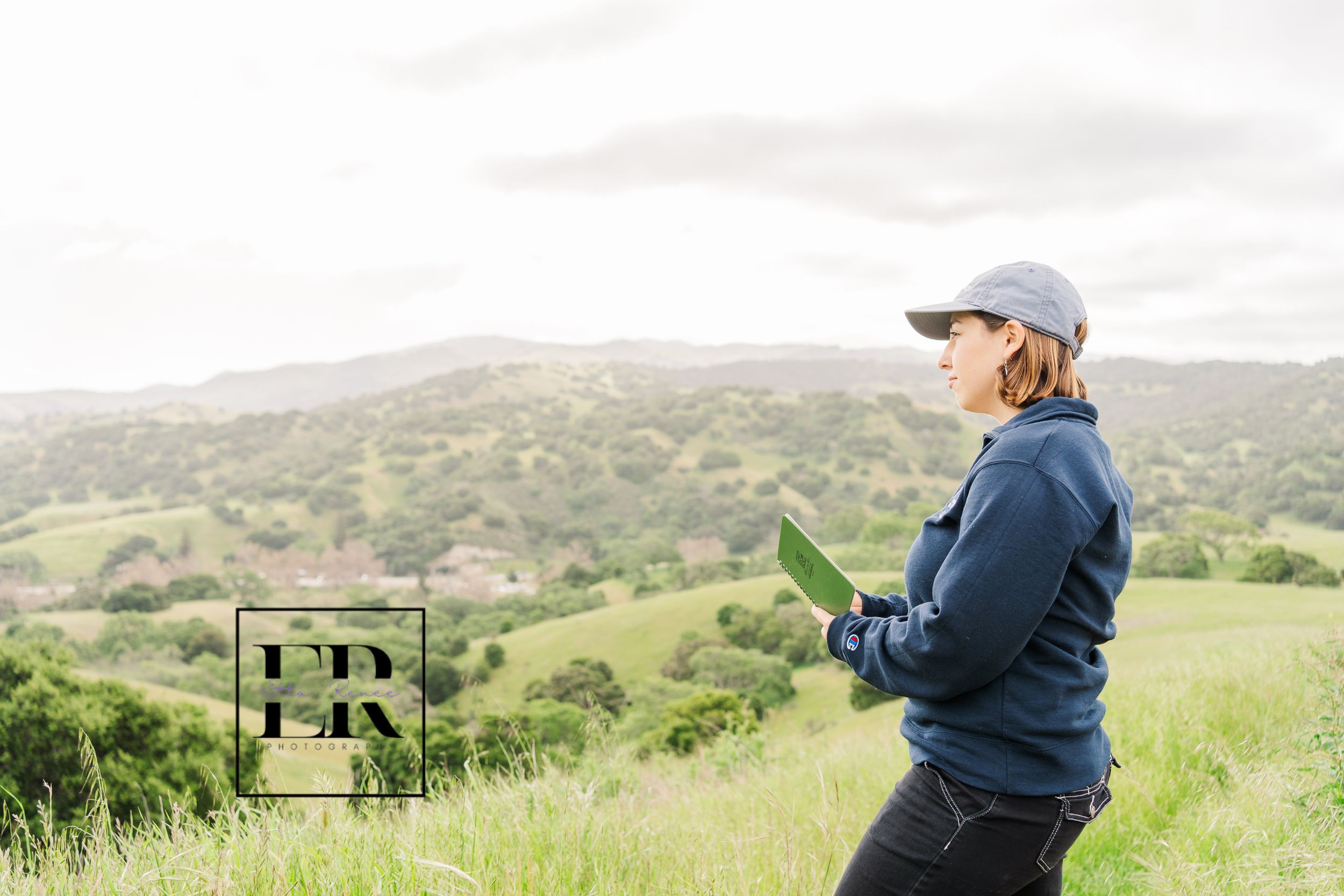 Hiker in navy jacket and cap explores rolling green hills while holding a mobile device on a cloudy day.