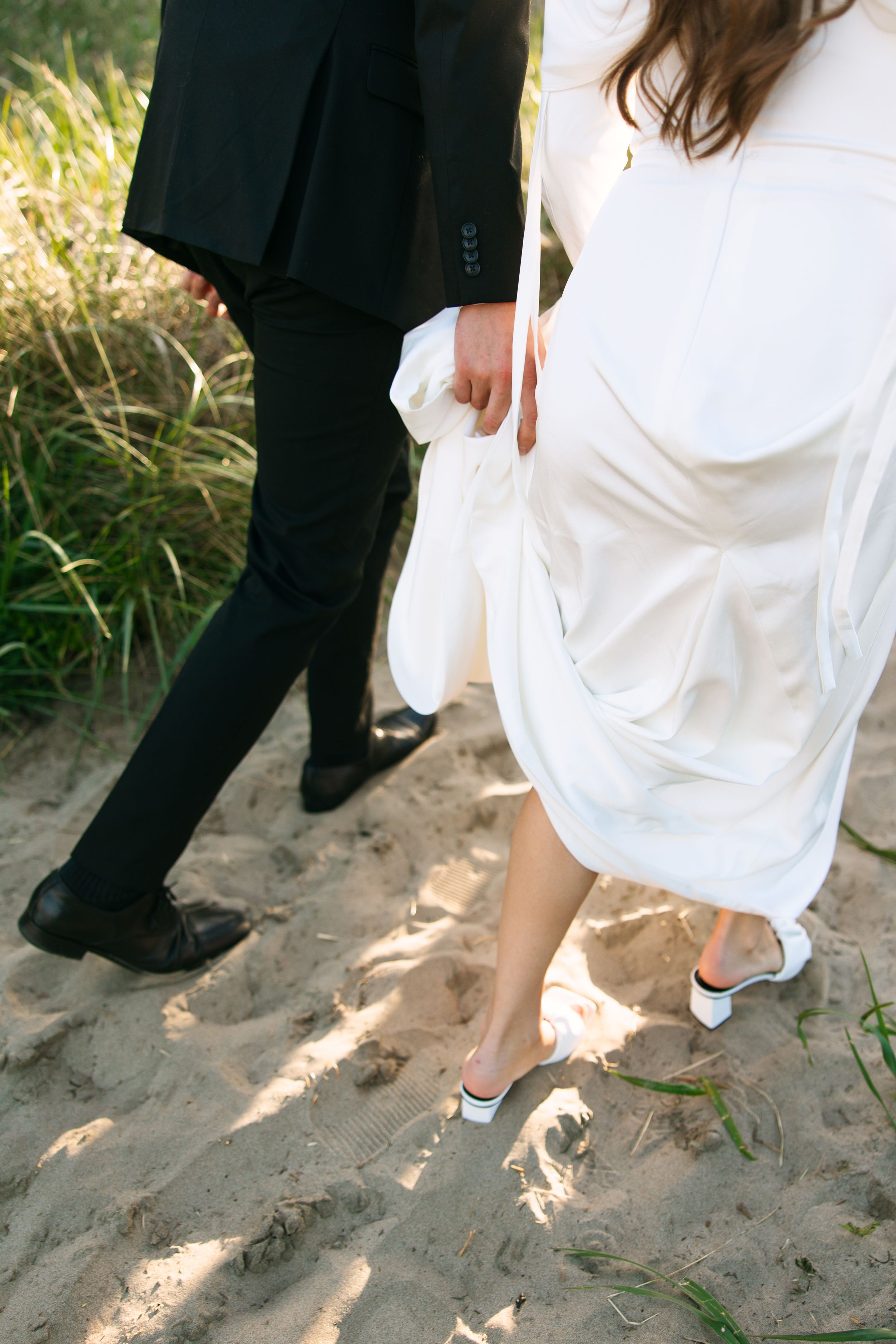 Close-up of bride and groom's feet walking on sandy beach, showing dress and formal shoes.