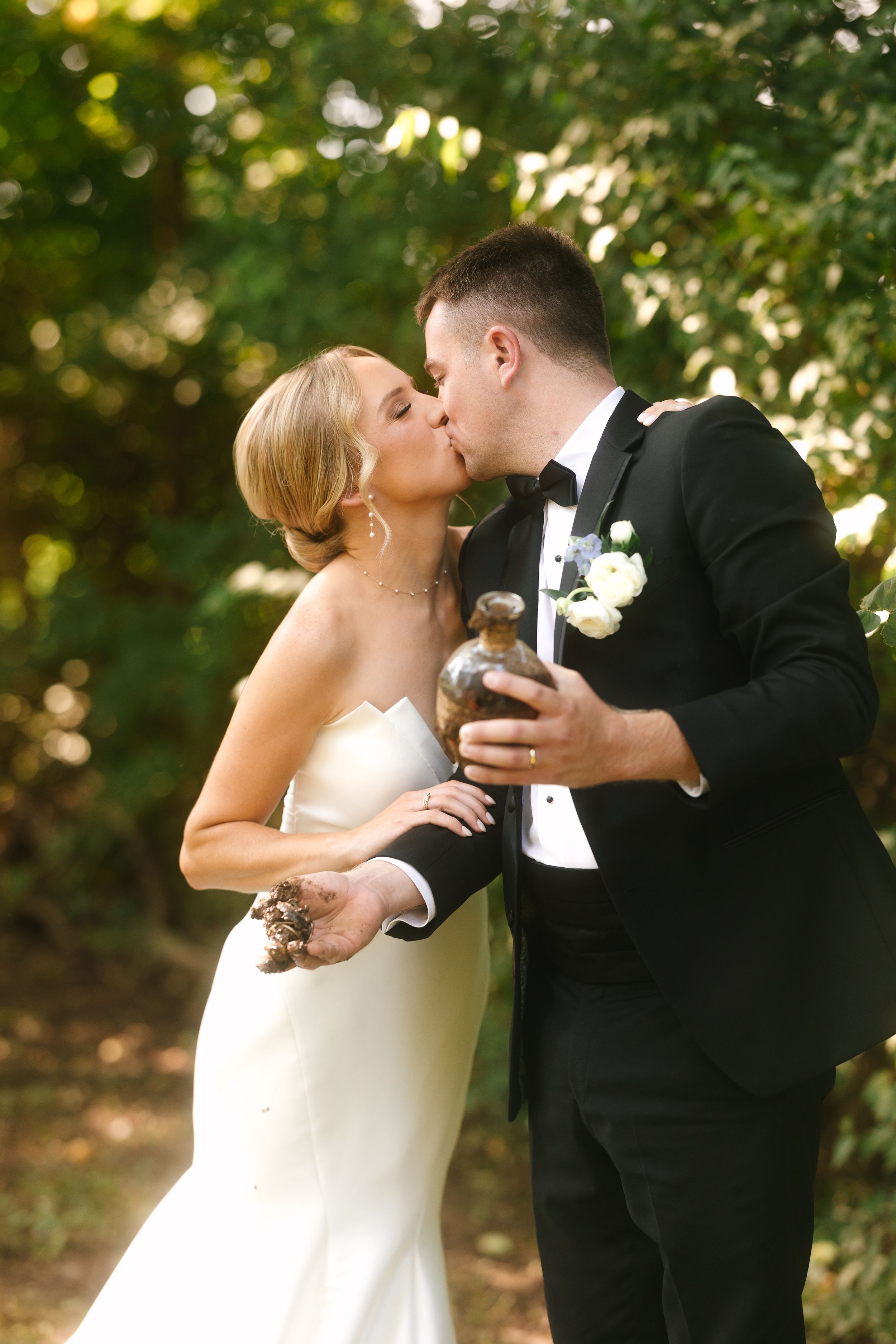 A couple shares a romantic kiss at their wedding celebration while holding drinks in an outdoor garden setting.