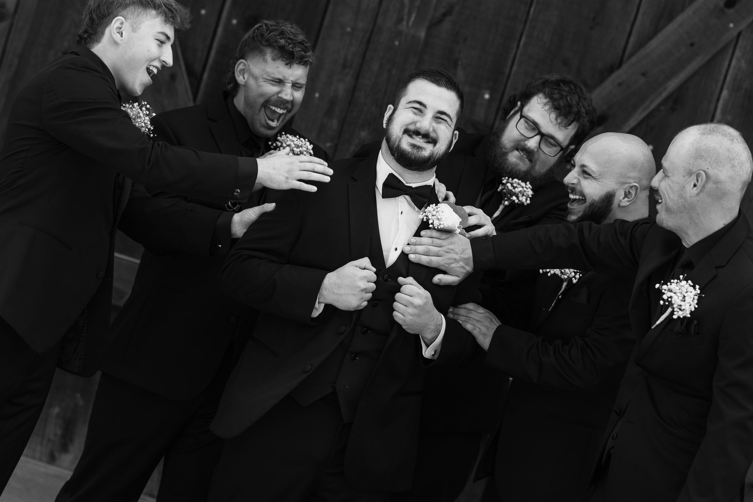 Black and white photo of groom laughing with groomsmen in suits, adjusting his bow tie before wedding ceremony.