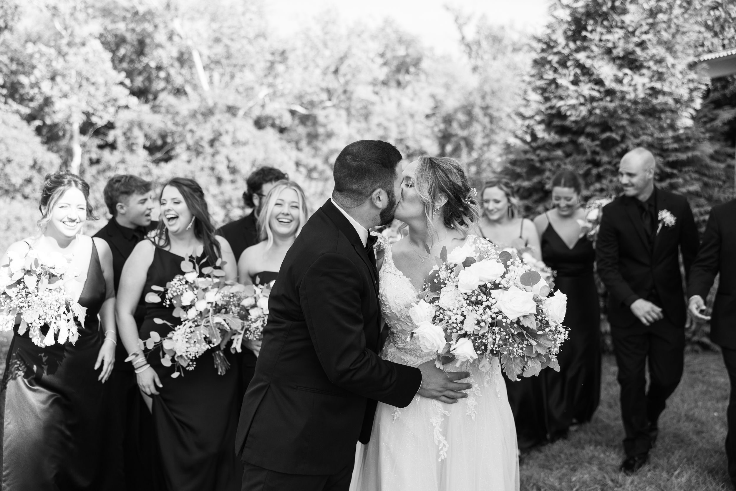 Black and white photo of a bride and groom kissing surrounded by their smiling wedding party outdoors.