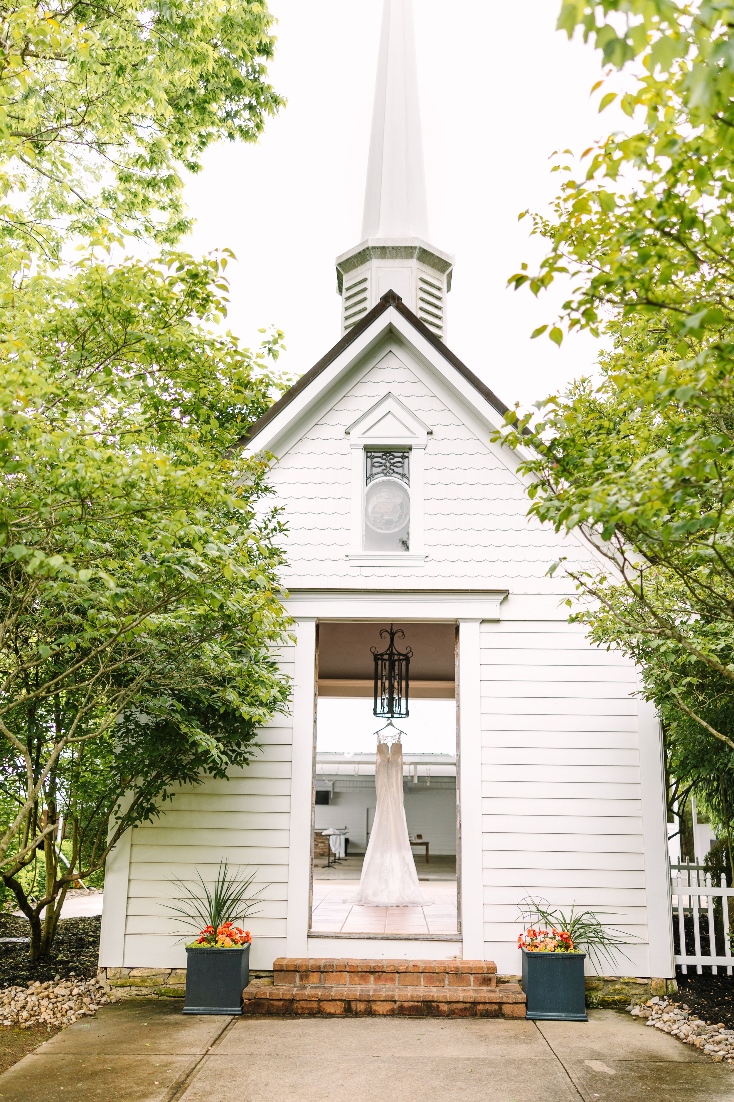 A charming white chapel with green trees and a wedding dress displayed in the doorway.