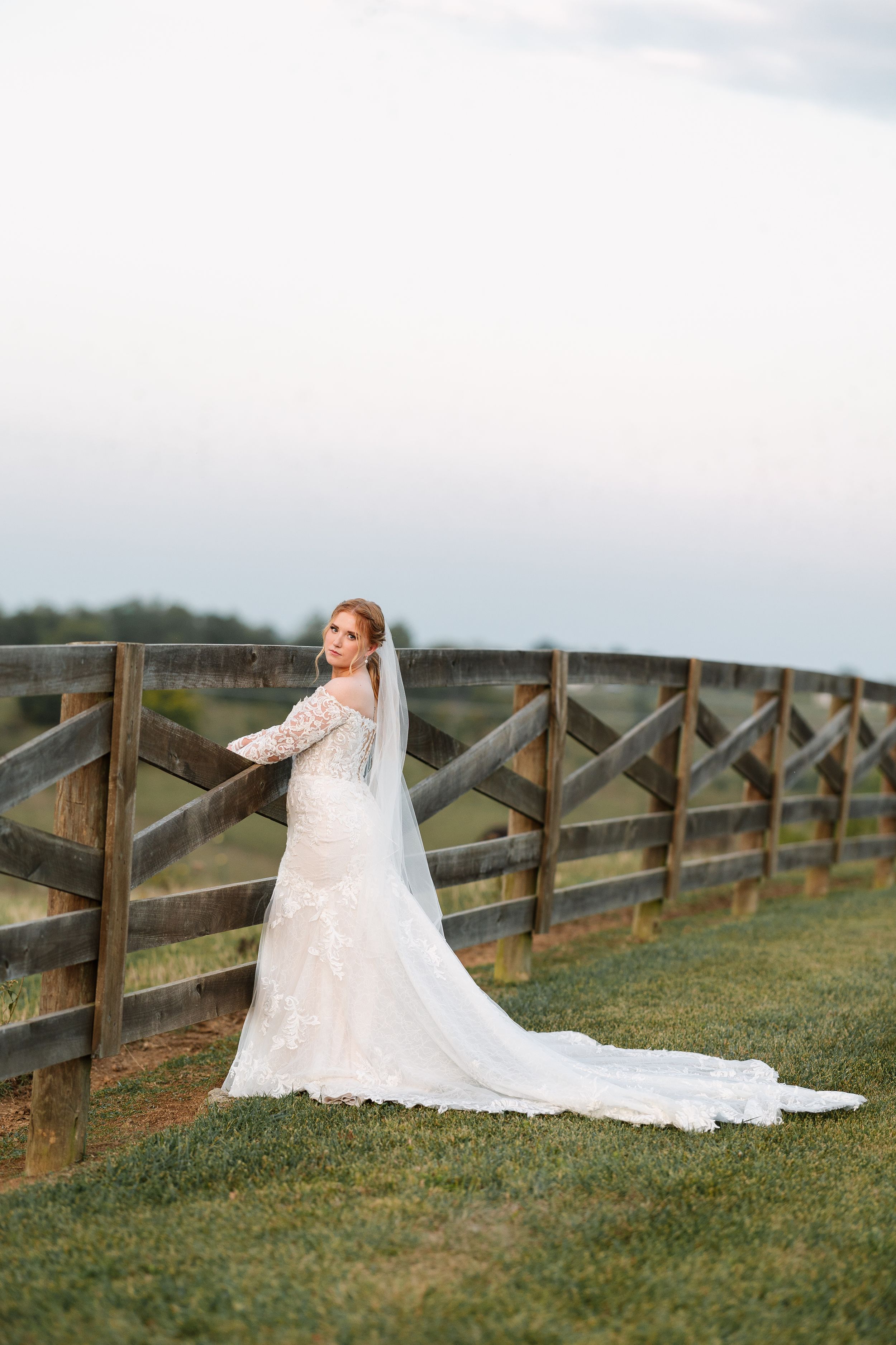 Bride in lace gown and veil poses by rustic wooden fence on green grass at outdoor wedding venue.