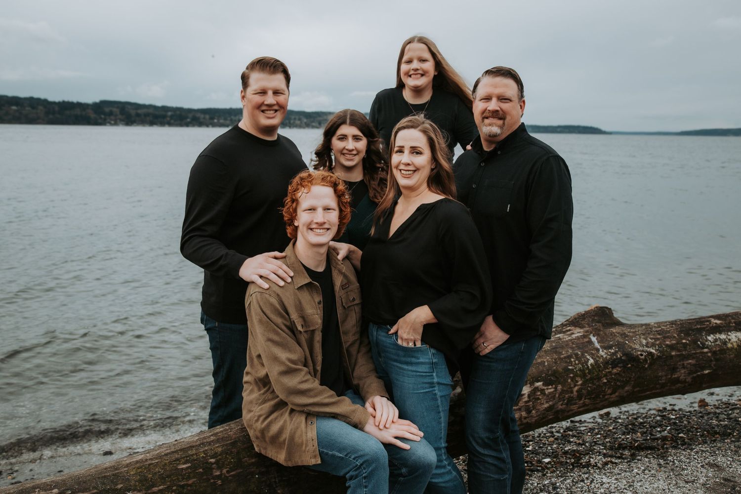 Family of six in dark clothing pose together on a driftwood log on a pebble beach with calm water and a forested shoreline behind.