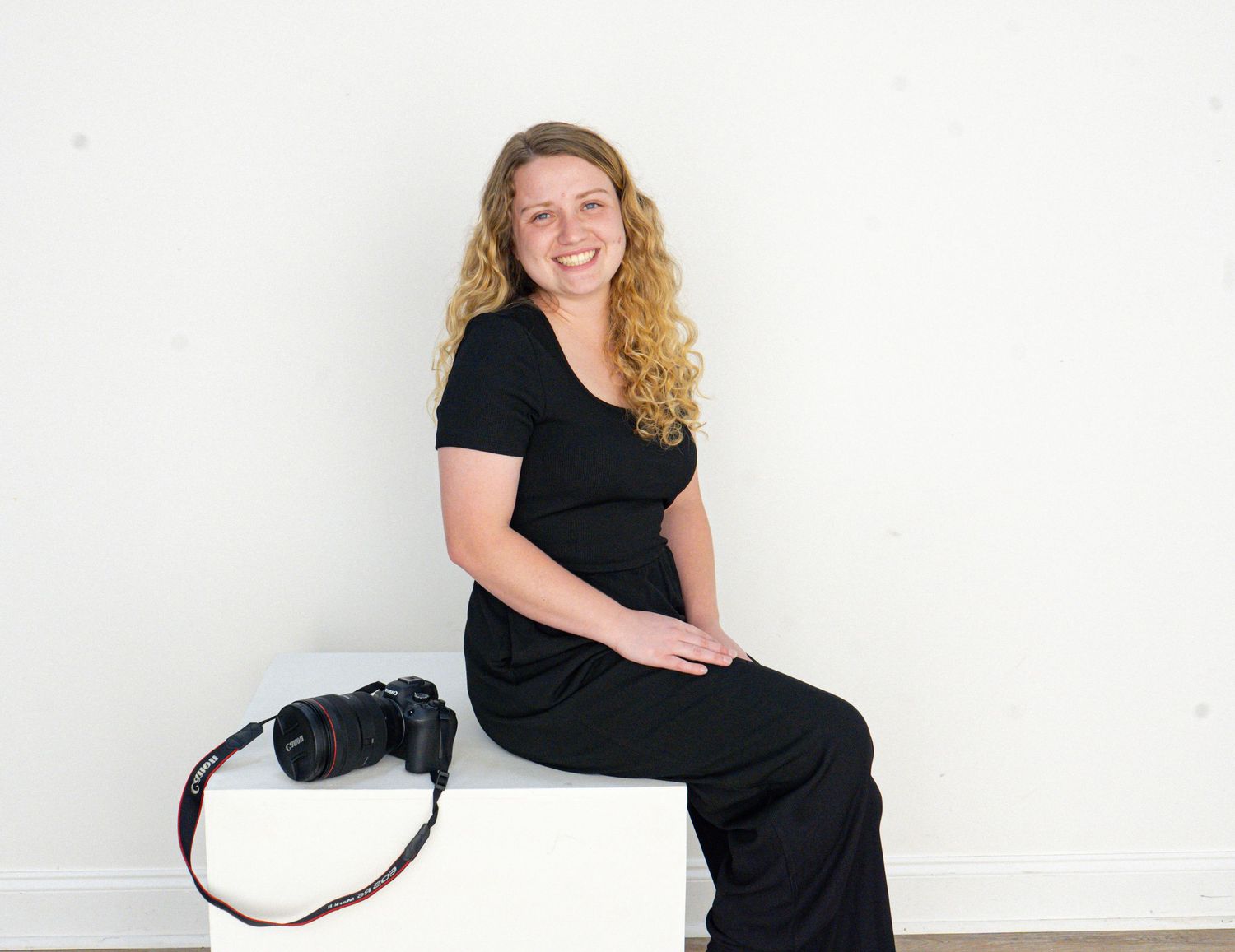Smiling female photographer in black outfit sitting with a mirrorless camera against a white background.