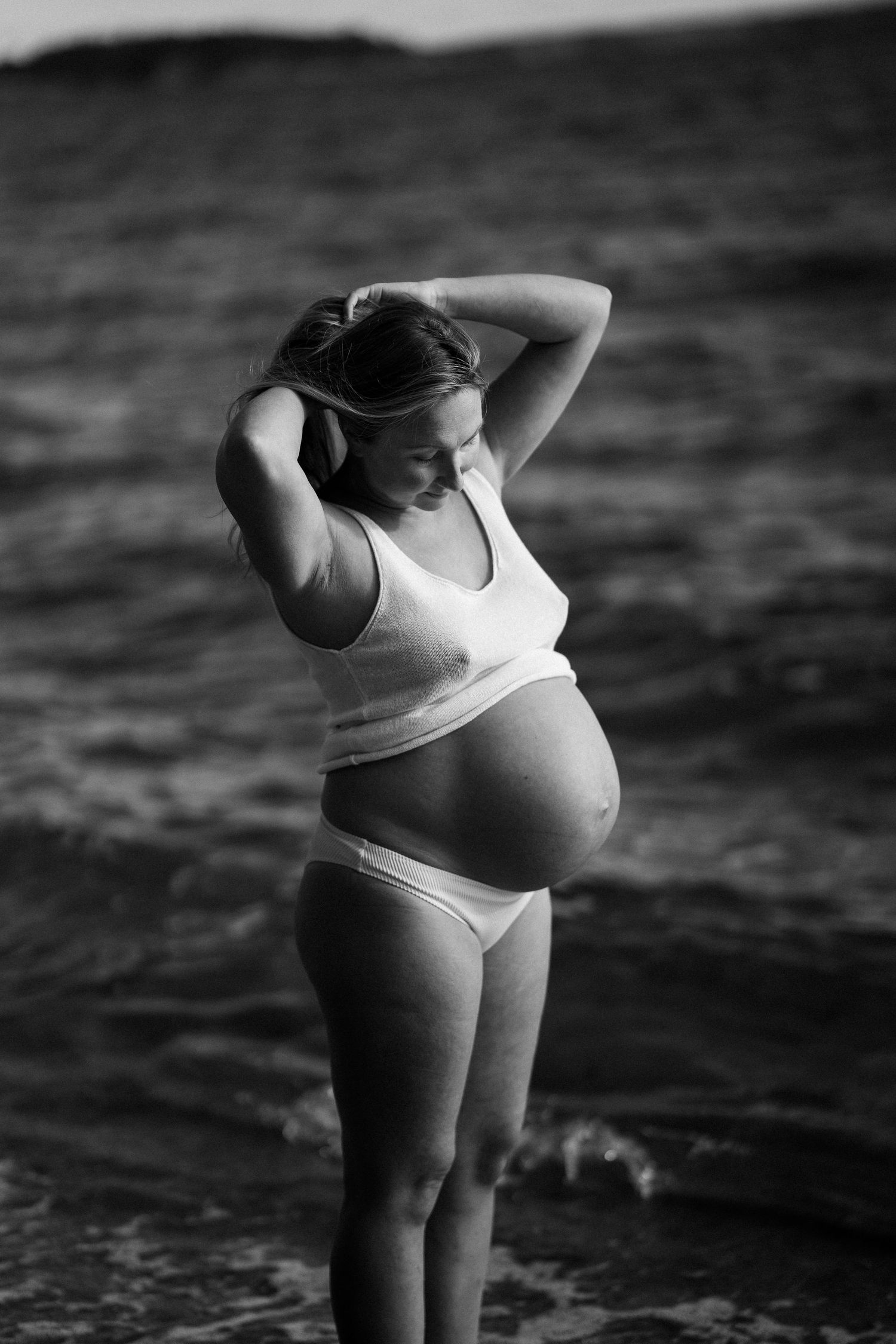 En gravid kvinna i vit topp och vita trosor står vid vatten och har händerna i håret.