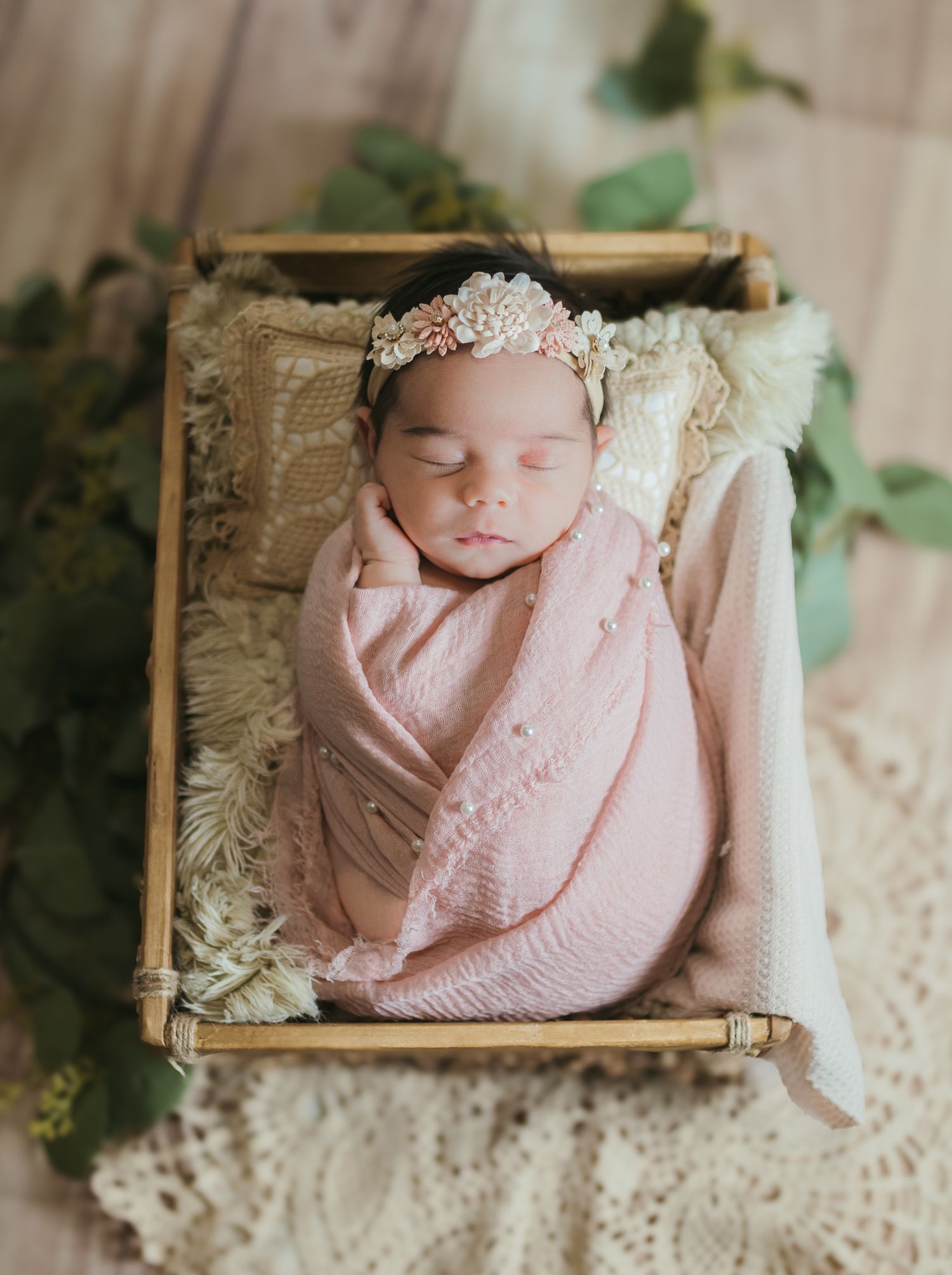 Newborn sleeping in pink swaddle with lace headpiece inside rustic wooden box with greenery.Colorado Springs Photographer