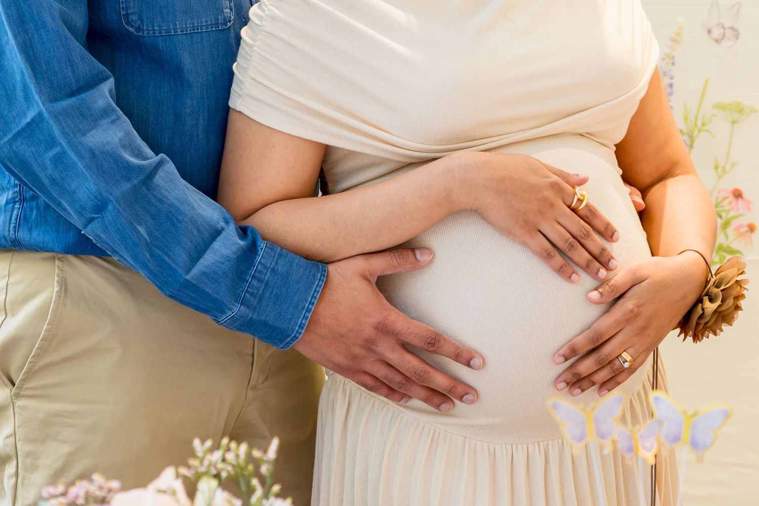 Close-up of pregnant couple cradling baby bump together, woman in white dress, floral background, maternity photo.