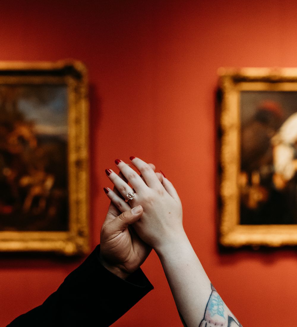 Couple holding hands with engagement ring against red gallery wall with gold framed paintings.