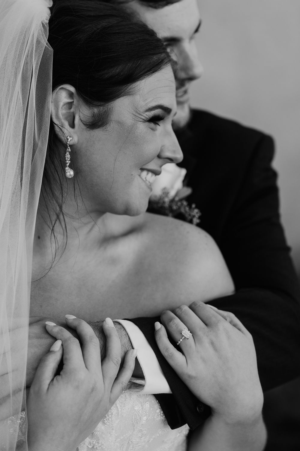 Black and white photo of smiling bride wearing veil and diamond ring embraced by groom.