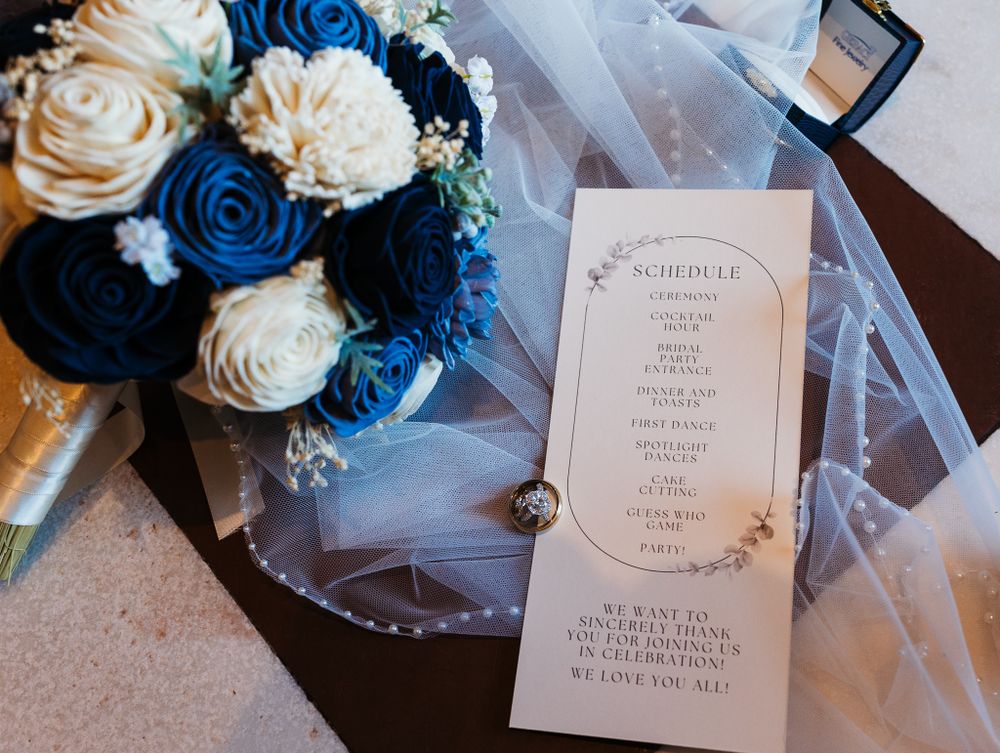 Wedding day flatlay with blue and cream rose bouquet, ceremony schedule program, veil, and diamond ring.