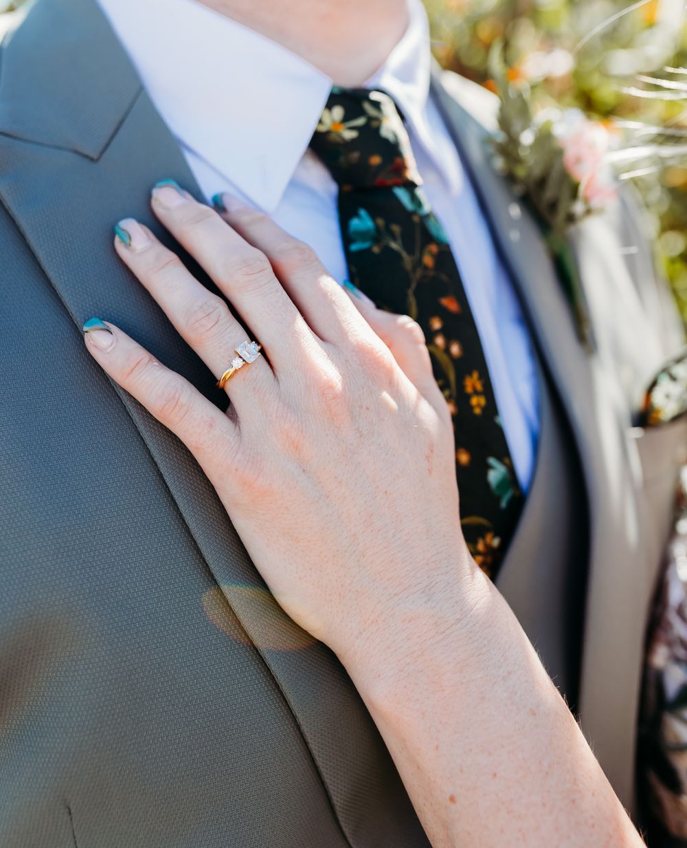 Bride's hand with diamond engagement ring resting on groom's grey suit with floral tie.