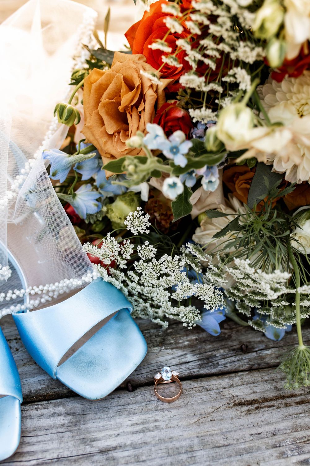 Bridal flat lay featuring blue satin heels, pearl-trimmed veil, diamond ring, and colorful bouquet with orange and red roses on wood.