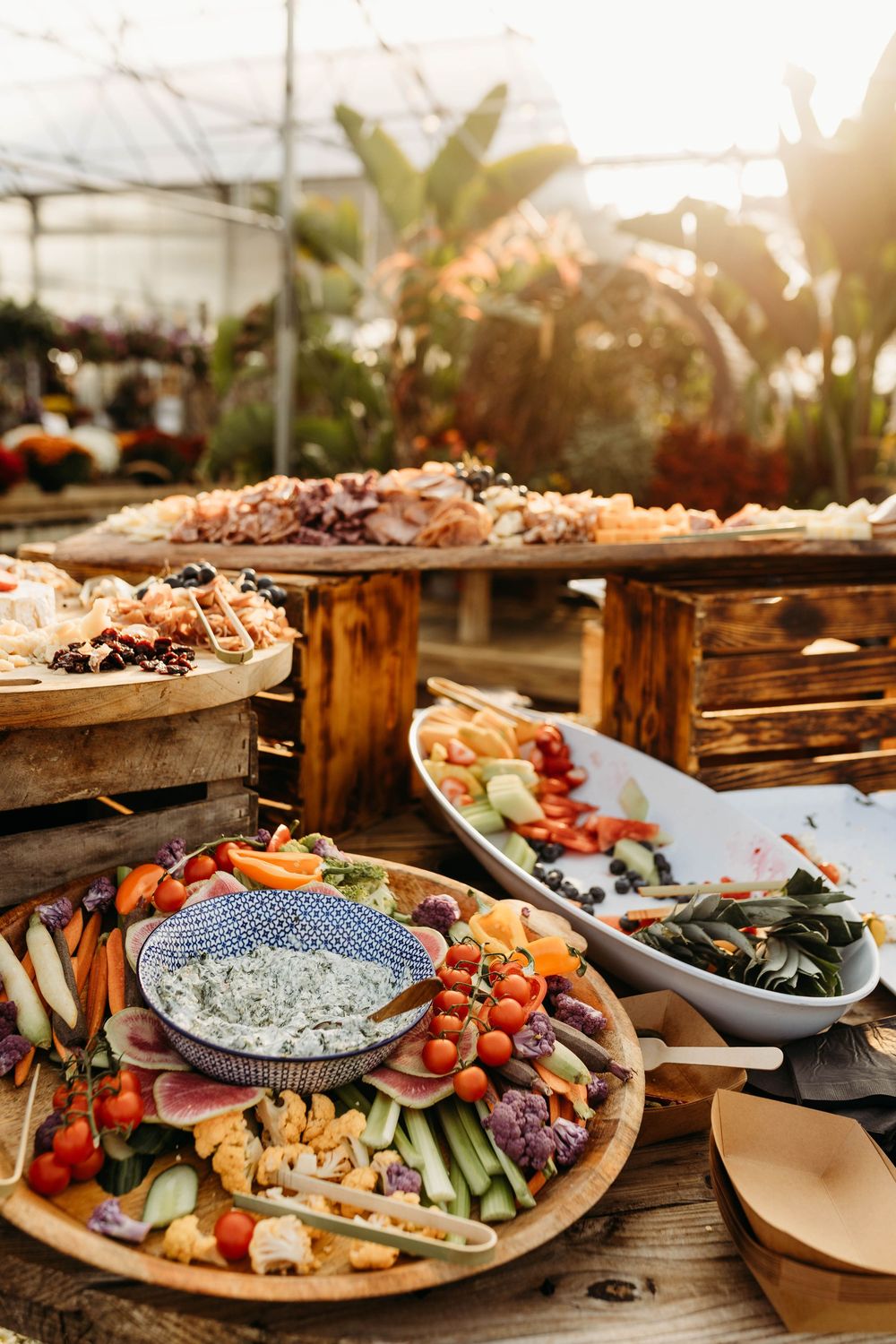 Abundant rustic wedding grazing table with charcuterie, vegetable crudites, dips, and assorted foods displayed in a sunlit greenhouse.
