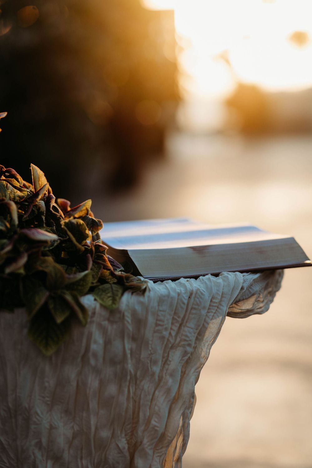 An open book rests on a white draped pedestal adorned with flowers during a golden sunset outdoor wedding ceremony.