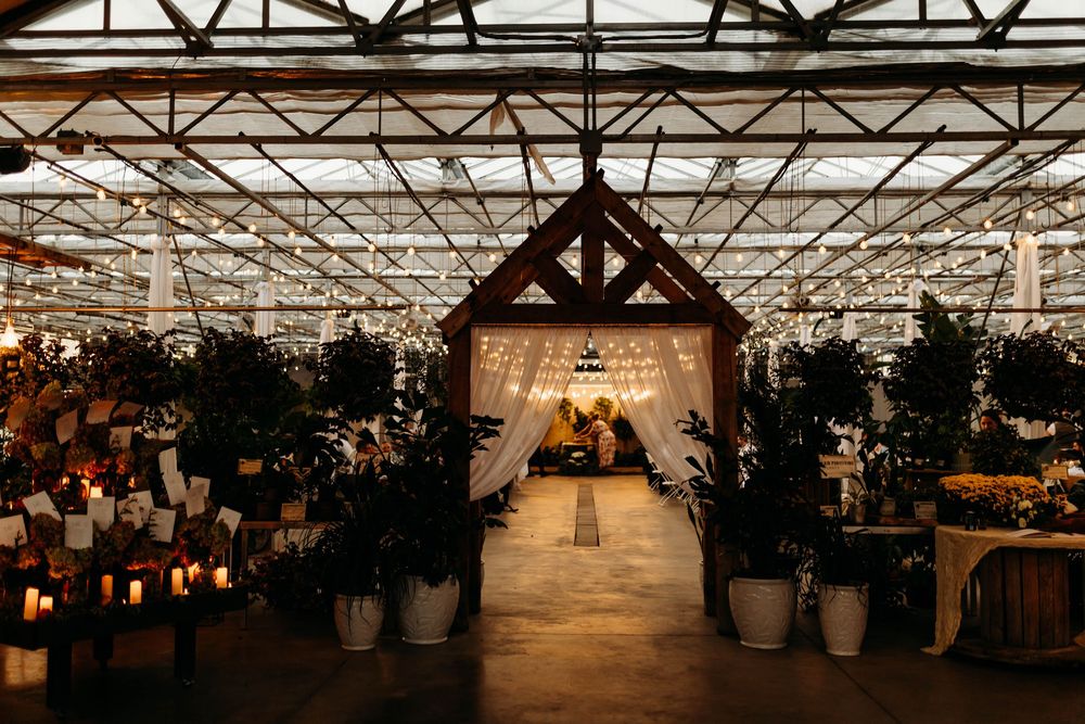 Elegant greenhouse wedding venue with string lights, candles, and lush plants lining a romantic aisle at dusk.