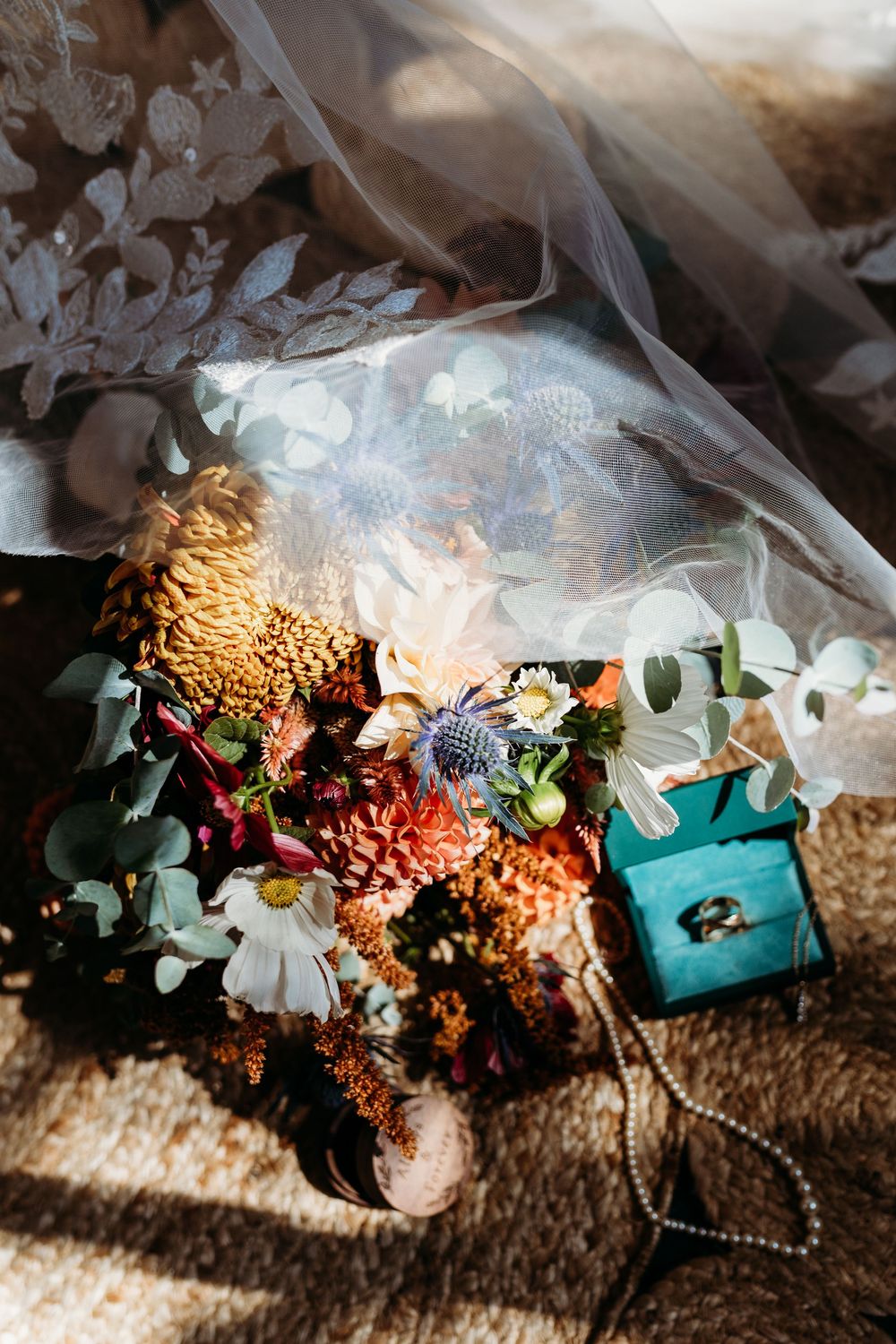 Bridal bouquet with colorful fall flowers beside a teal ring box with wedding bands under a lace veil.