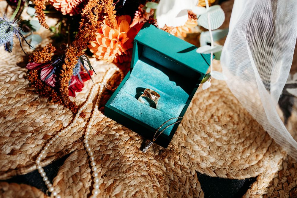 Gold wedding bands displayed in a teal velvet ring box beside a colorful bridal bouquet on woven texture.