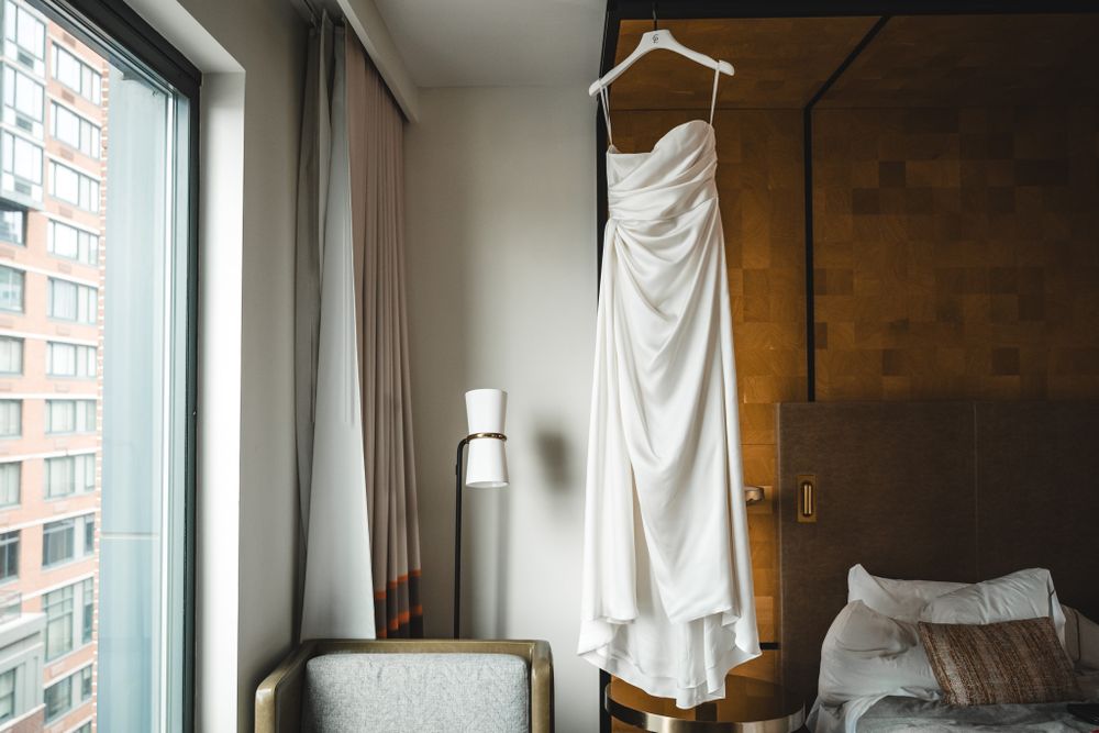 Wedding dress hanging in window of modern hotel room with city view.
