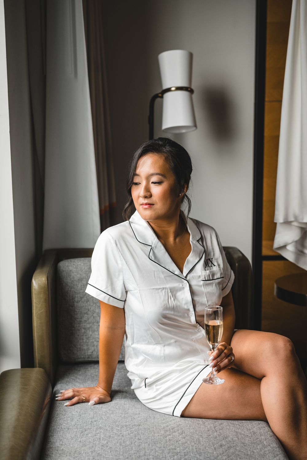 Person in white pajamas relaxing with wine on gray couch near window.