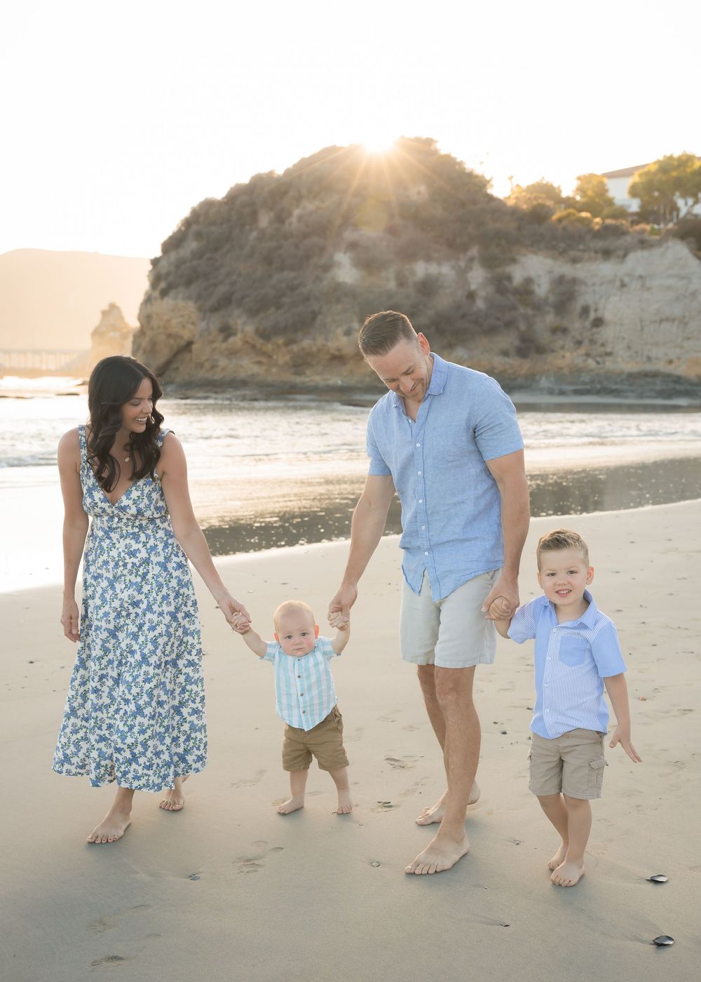 Family of four walking barefoot on a sunny beach at golden hour, with rocky cliffs in the background.