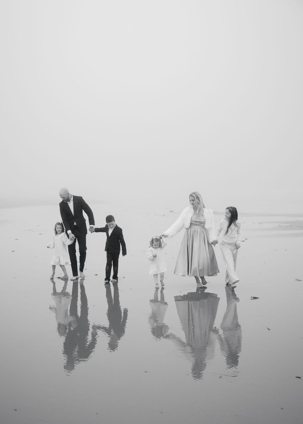 A family group stands on a reflective beach in black and white, creating mirror-like silhouettes in the wet sand.